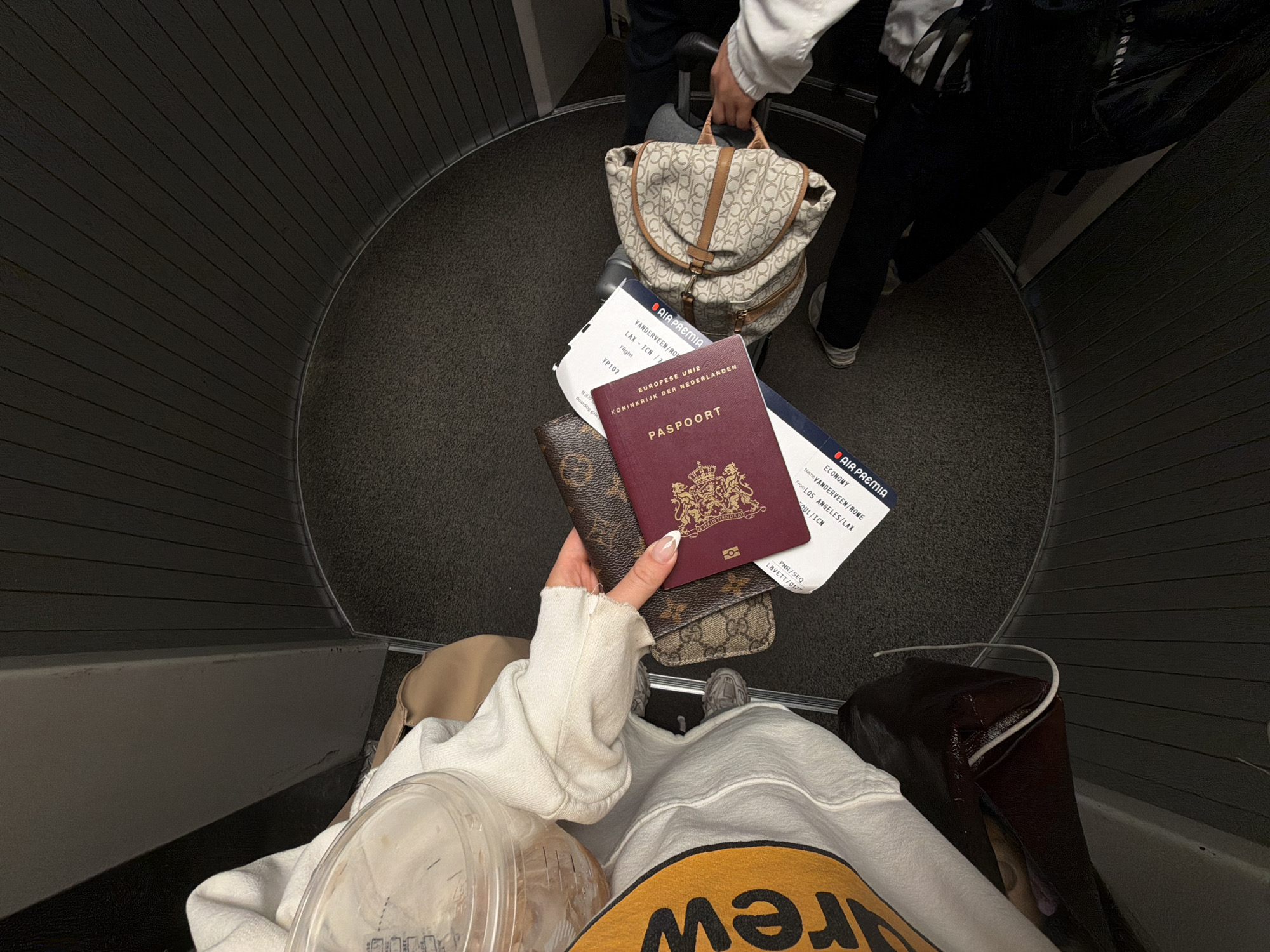 The image, captured from a high angle looking down, shows a person in a dark, curved hallway, likely an airplane boarding bridge. The primary subject's torso, arms, and legs are visible. They are wearing a white long-sleeved top with distressed cuffs, a yellow garment featuring the "DREW" logo underneath, and grey sneakers. In their hands, they hold a burgundy Dutch passport, prominently displaying "PASPOORT," "EUROPESE UNIE," and "KONINKRIJK DER NEDERLANDEN." Accompanying the passport are a Louis Vuitton monogrammed passport holder and a partially visible Gucci monogrammed card holder. An Air Premia boarding pass is also held, showing the name "VANDERVEEN/ROME," flight "YP102," "ECONOMY" class, and the route "LOS ANGELES/LAX" to "SOUL/ICN." A clear plastic cup with a lid is also in their grasp.
Another person stands further into the hallway, partially visible, holding a light-colored backpack with a "CK" monogram pattern and brown leather straps, which rests on what appears to be rolling luggage. This person wears a white top and dark pants. The floor of the hallway is covered in a dark gray carpet, while the walls are dark with horizontal grooved panels, contributing to the enclosed and transitional setting. The scene depicts individuals in transit, indicating air travel and possibly boarding or disembarking a flight from Los Angeles.