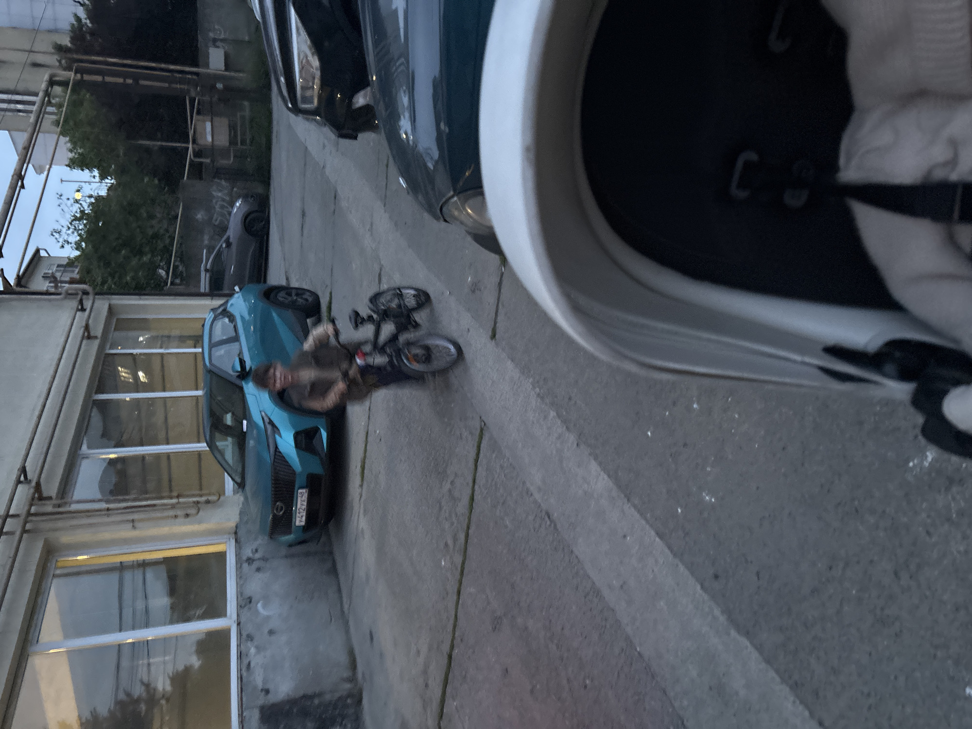 The image appears to be taken from a first-person perspective, possibly from inside a vehicle. A partially visible white helmet is in the foreground.
The scene is an outdoor environment in what seems to be an urban or industrial area. A blue car is parked near a building with large windows. A person is sitting on a small bicycle next to the car. Several other cars are parked nearby. The license plate on the blue car has a "48" code, indicating that it is from the Lipetsk region in Russia. The location, based on this information and the overall appearance of the environment, could potentially be Sochi, Russia.