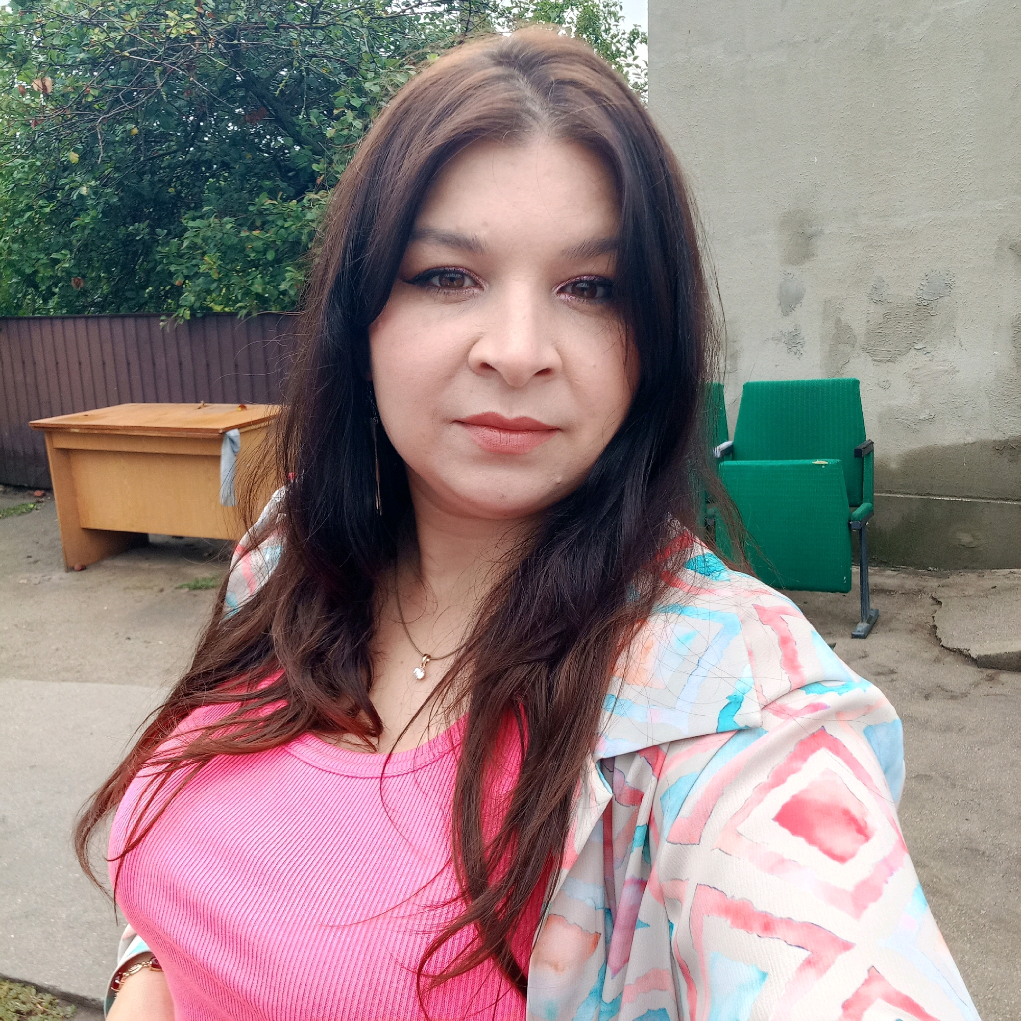 A woman with long, dark brown hair and brown eyes, wearing a pink ribbed tank top and a colorful jacket, stands outdoors in front of a building. Behind her, to the left, is a wooden desk and a wooden fence. To her right are two green chairs and a concrete wall. The ground is paved. The woman is looking directly at the camera, with a slight smile. The scene appears to be in a residential or industrial area.