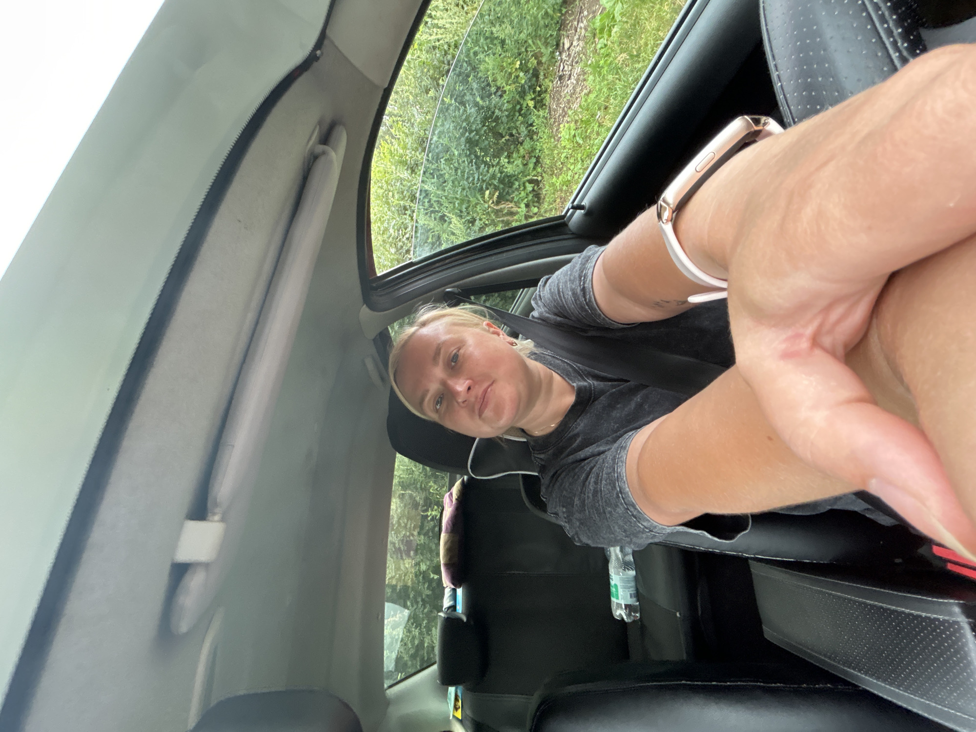 A woman is pictured inside a car, possibly in Chaikovsky, Russia, based on the provided context. She is in the driver's seat, with her right arm extended towards the camera. She is wearing a dark-colored t-shirt and a watch on her wrist. The interior of the car is visible, including the headliner, sun visor, seats, and a water bottle on the back seat. Outside the car window, greenery and trees are visible, suggesting a natural or rural setting. The woman is looking towards the camera with a neutral expression.
