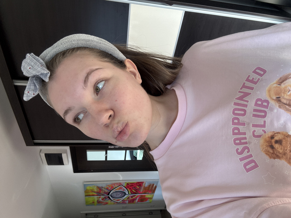 The image depicts a young adult female in an indoor residential-like setting, located in Vozdovac Urban Municipality, Serbia. The female is positioned from the mid-chest upwards, angled slightly to her left, with her head tilted back and eyes looking upwards and to her left. Her lips are pursed in a "duck face" expression. She has fair skin, light brown hair, and appears to be wearing no visible makeup. A small mole is present on her left cheek, below her mouth. She wears a grey, terry-cloth textured headband with a small plaid bow (grey with red dots) atop her head, holding her long, light brown hair back. She is dressed in a loose-fitting, light pink crew-neck t-shirt. The visible print on the shirt reads "DISAPPOINTED CLUB" in pink text, with the upper portions of two animal images below it: a light brown fluffy dog on the left and a brown lop-eared rabbit on the right.
The background reveals a white wall on the left side, featuring a vertical abstract painting. This artwork showcases a central eye-like motif within a white circle, surrounded by radiating lines and splatters of yellow, red, and orange. To the right of the painting, a dark wooden door with a rectangular frosted glass panel is partially visible. Above this door, a white rectangular unit, possibly an electrical box or intercom, is mounted on the wall. The right side of the background is dominated by a dark brown wardrobe or closet with sliding doors, one of which features a white panel on its lower section.