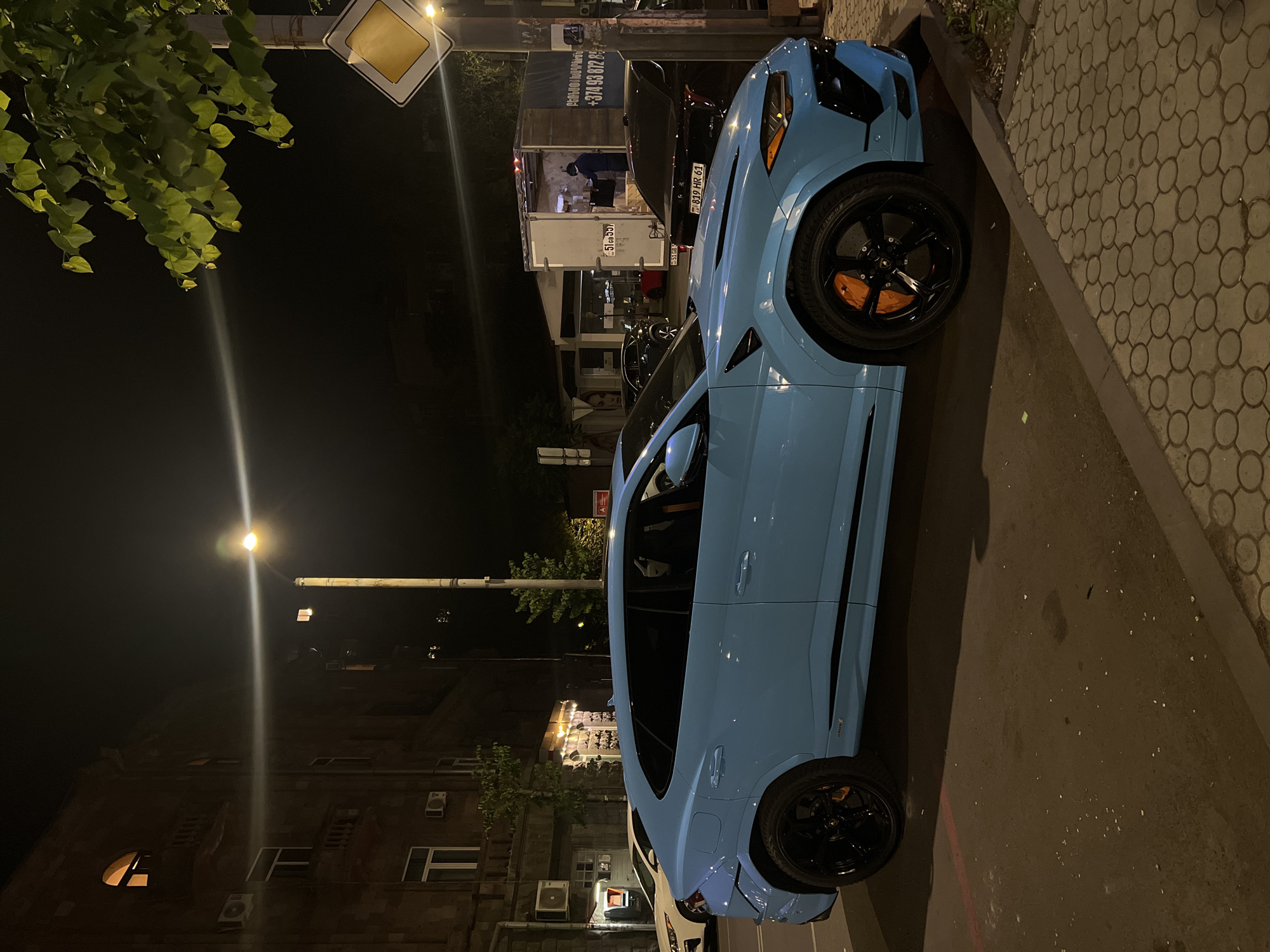A light blue Lamborghini Urus is parked on a street in Yerevan, Armenia at night. The car is facing the left of the frame. The street is paved with cobblestones on the right side and asphalt on the left. There is a building on the left side of the frame, an apartment building with multiple windows and air conditioning units. A street light illuminates the scene. On the right side of the frame, there is a yellow diamond-shaped road sign, and a white box truck is parked, with a person standing in the open back of the truck. The license plate of the truck is visible, reading "51 GB 337". Another black car is parked behind the truck, with a license plate reading "819 HR 61". A tree with green leaves hangs over the right side of the frame. The scene is dimly lit by streetlights and the interior lights of the vehicles and buildings.