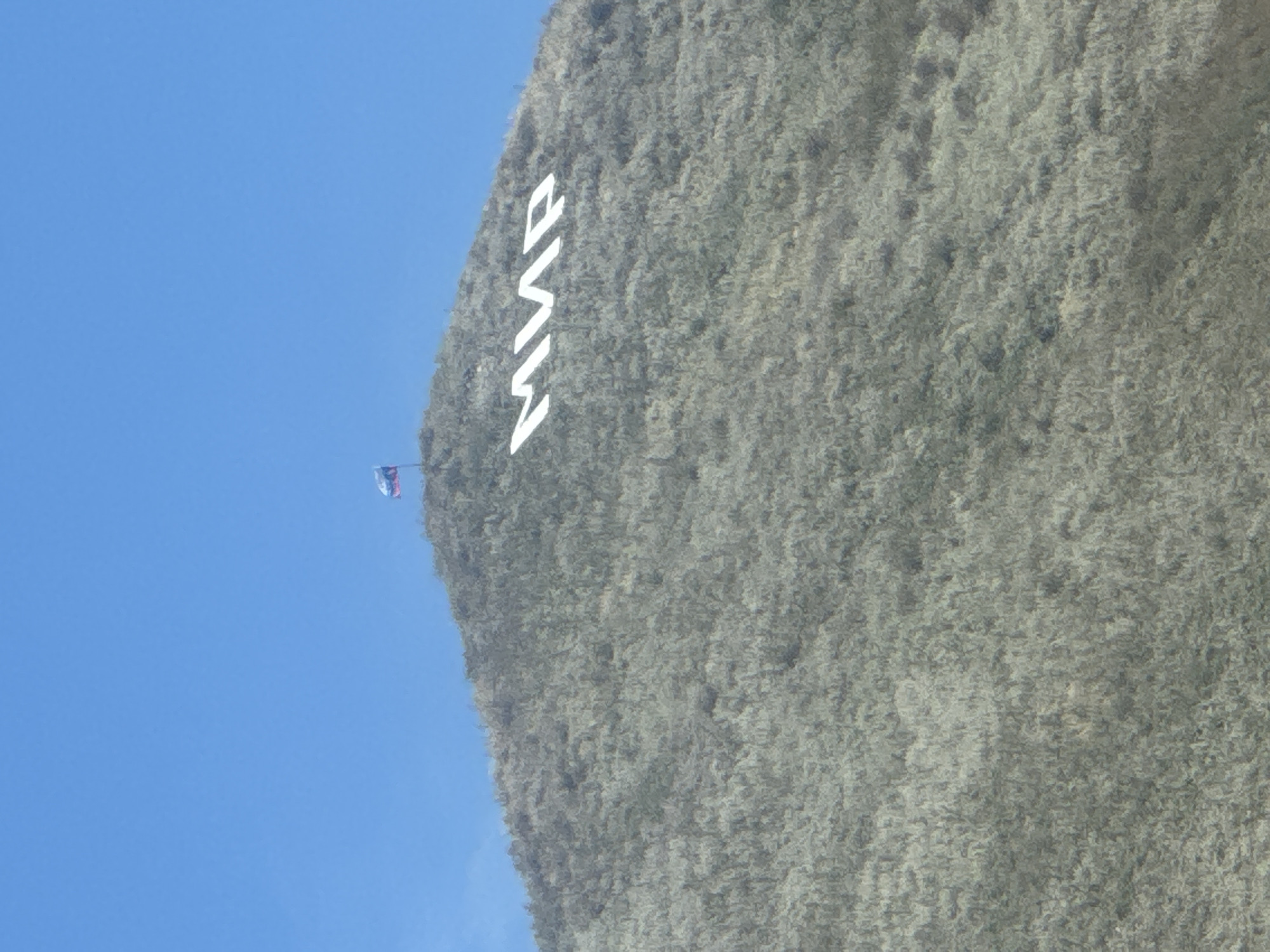 The image shows a wide shot of a grassy hill with white lettering spelling "МИР" on its side. A small Russian flag flies from a pole near the top of the hill. The sky above is clear and blue. The location is the hill in the city of Кугарчи, Russia, which features the lettering "МИР" (meaning "peace" in Russian) as a prominent landmark.