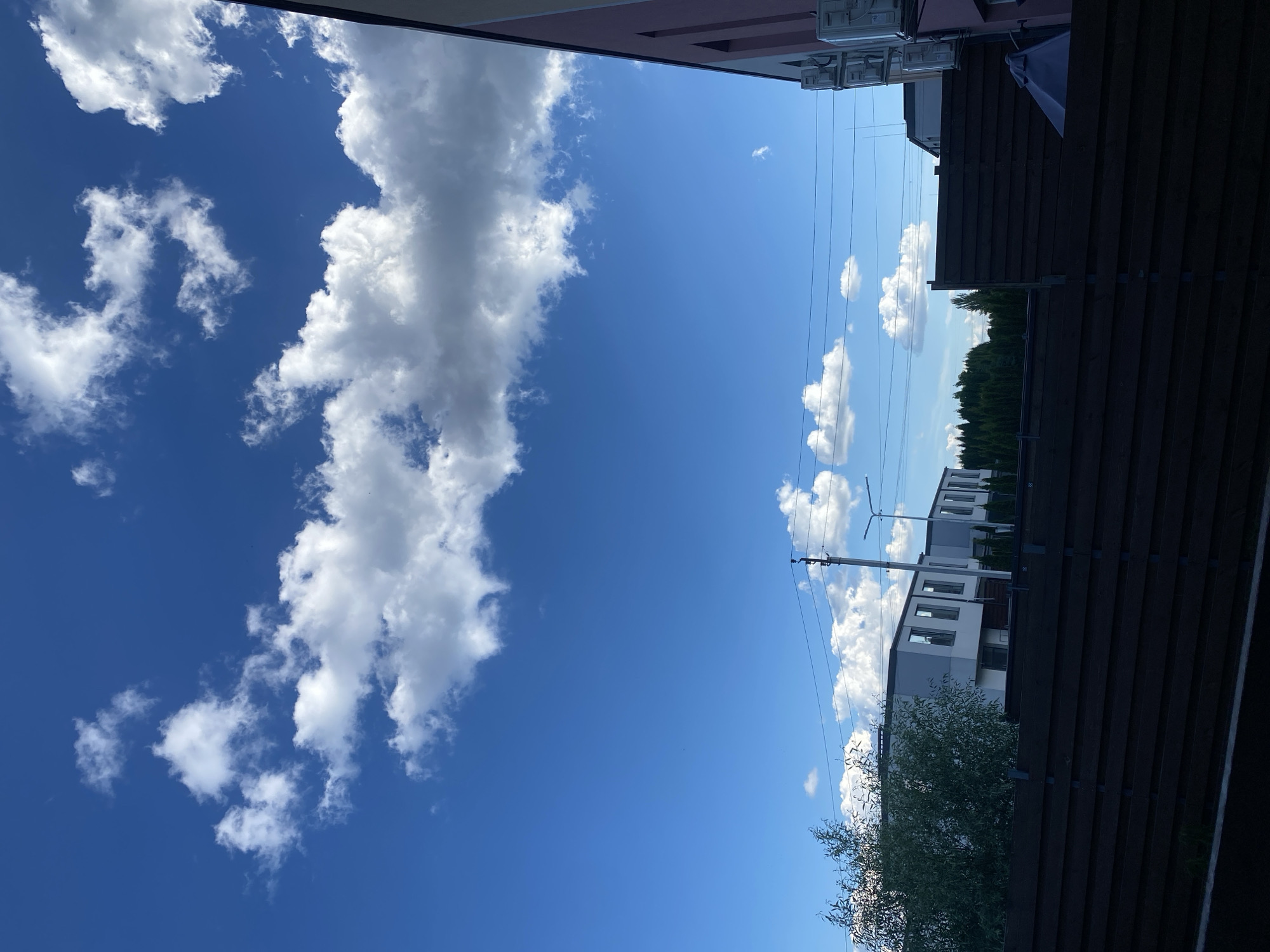 The image captures a daytime exterior scene in Novi Petrivtsi, Ukraine. The sky is predominantly blue with scattered white cumulus clouds. In the foreground, a dark wooden fence stretches across the lower portion of the frame. Behind the fence, several modern houses with light-colored facades and dark-framed windows are visible. A line of green coniferous trees forms a backdrop behind the houses. Electrical wires and a utility pole with streetlights are also present in the middle ground. The right side of the image shows the corner of a building with a pink and gray exterior, featuring multiple outdoor air conditioning units mounted on the wall. There are no people or animals visible in the scene.
