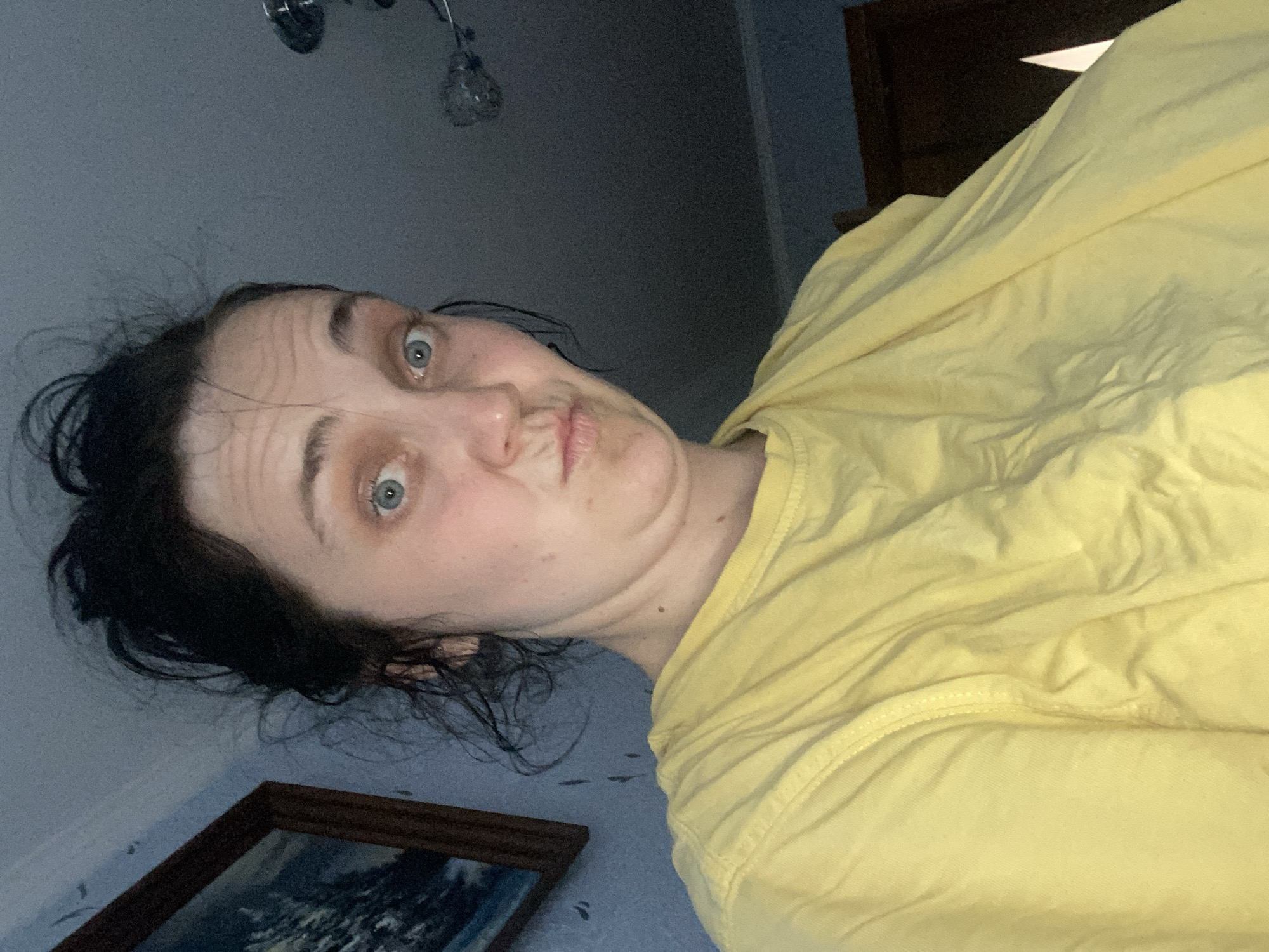 A woman with dark, messy hair and blue eyes, wearing a yellow t-shirt, makes a duck-face at the camera. She is indoors, in what appears to be a bedroom. A framed picture hangs on the wall behind her, and a ceiling fan is visible in the upper right corner. The room is dimly lit, with light coming from the ceiling fan.