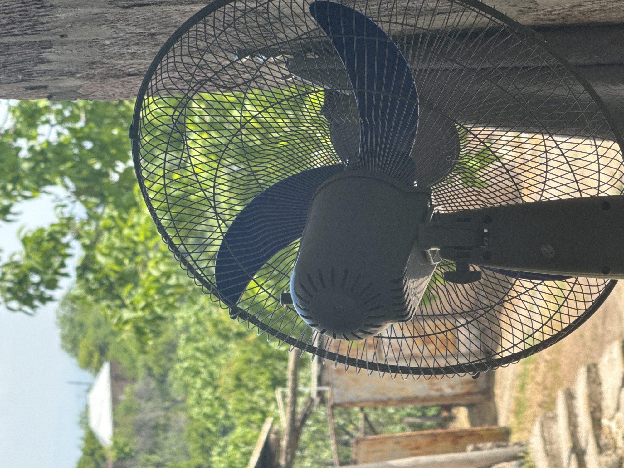 A close-up, outdoor vertical photograph focuses on a dark gray electric pedestal fan positioned in the foreground, with its back visible and angled slightly towards the left. The fan features a dark blue, three-bladed rotor enclosed within a black metal safety grille with a diamond mesh pattern. A black control knob is located on the top of the fan's motor housing, and a 'QC PASS' sticker is affixed to the lower portion of its stand on the right side. The background is softly blurred, revealing a verdant, sunlit landscape dominated by lush green trees and foliage. To the far left, partially visible rustic structures, possibly involving wooden posts and corrugated metal sheets, are present. In the distant center-left, a building with a light-colored, peaked roof is partially visible amidst the dense greenery. The ground to the left of the fan consists of dry earth and irregular light-colored stone or concrete steps leading downwards. To the right, a rough, textured vertical surface, resembling a tree trunk or weathered wooden wall, partially frames the scene. No individuals are present. The location is in the city of Unknown, Unknown.