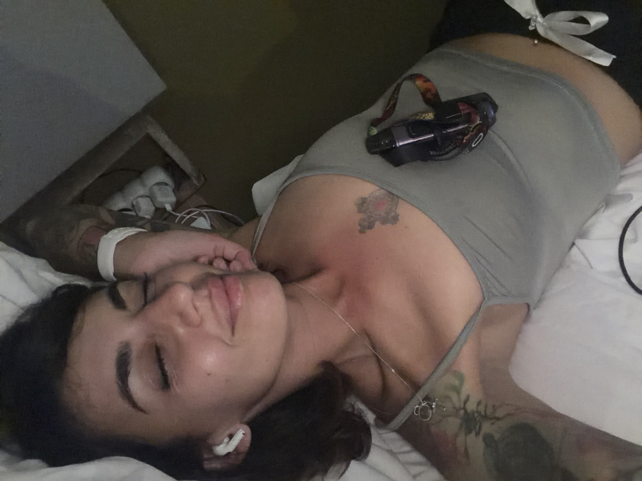 This image depicts a female individual lying on white bedding in an indoor setting, identified as Nusa Dua, Indonesia. She is positioned diagonally in the frame, appearing to be resting or asleep with her eyes closed and a slight smile. Her hair is dark and of medium length. She wears a light grey or greenish crop top, displaying a small cross-like tattoo on her upper right chest and extensive sleeve tattoos on both arms. A white wireless earbud is in her left ear, and a thin silver necklace with a small pendant is on her neck. A white wristband is present on her right wrist. A black vaping device with a colorful lanyard is tucked into the left side of her crop top. Her navel piercing with a small stud is visible, along with a portion of black bottoms featuring a white bow. In the upper right background, a dark olive-green wall with a wooden frame and multiple white electrical outlets and wires is visible.