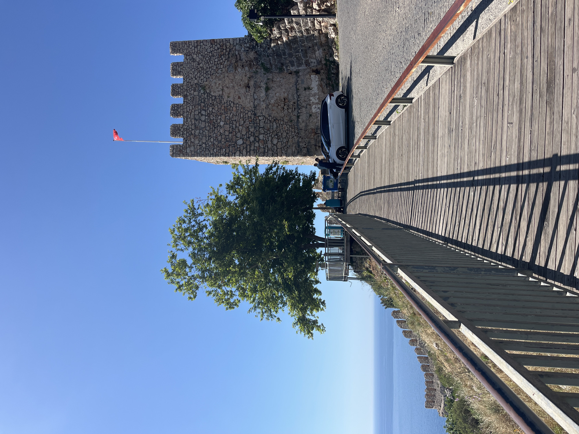 The image captures a sunny day in Alanya, Turkey, with a view that includes historical elements and the Mediterranean Sea. An old stone castle tower with battlements stands prominently, topped with a red flag. Adjacent to the tower, a paved road leads onto a wooden boardwalk with a railing. A white car is parked alongside the tower. Below the boardwalk, a cliff edge is visible with stone ruins overlooking the sea. A large tree with green foliage partially obscures the view of a lookout point. The sky is clear and blue, contributing to the bright, outdoor scene.