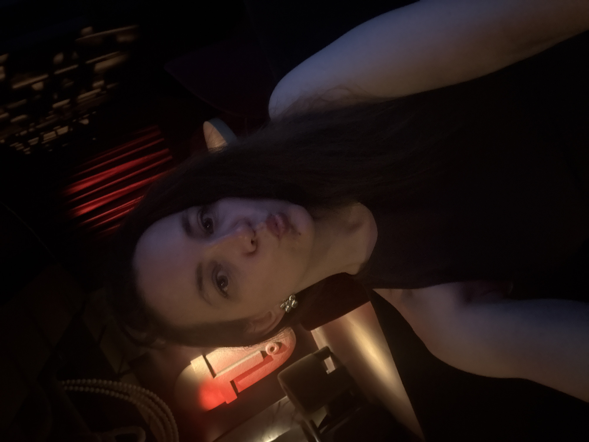 The image shows a person with long brown hair and fair skin lying down, possibly on a couch or bed. The person is wearing a dark-colored top. The environment appears dark, with red light reflecting off of the ceiling. There are some objects in the foreground, including what looks like a black choker with a charm. Due to the low light conditions and lack of landmarks, it is impossible to determine if this is in Moscow, Russia.