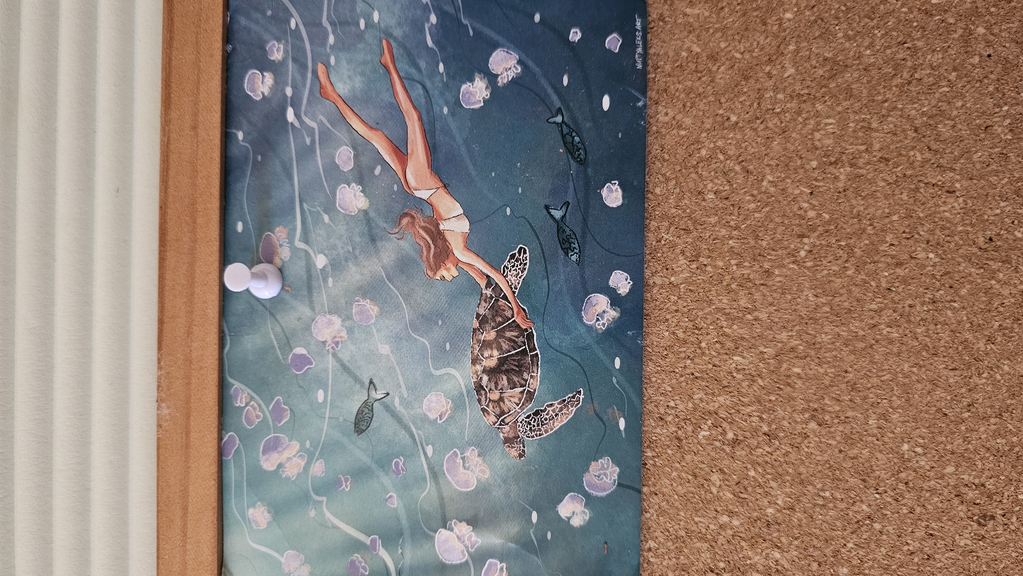 The media file shows a piece of art, possibly a print or illustration, depicting a woman swimming with a sea turtle. The scene is underwater, suggested by the blue-green hue of the background and the presence of jellyfish and small fish. The woman is wearing a white bikini and her body is extended as she gracefully interacts with the turtle, holding onto its shell. The turtle is large and detailed, with a patterned shell. Both subjects are rendered in a slightly stylized, illustrative manner.
The artwork is pinned to a corkboard, with a wooden frame visible on the left side. A white pushpin is used to secure the artwork. The lighting appears to be from an overhead source, casting subtle shadows.
The visible text in the upper right corner of the artwork reads "MATHILDE ART".
The overall mood of the image is serene and evokes a sense of harmony with nature. There are no strong cues for time of day or weather beyond the underwater setting. The location context provided, Baan Chaweng, Thailand, is a coastal area known for its beaches and marine life, which aligns with the artwork's theme.