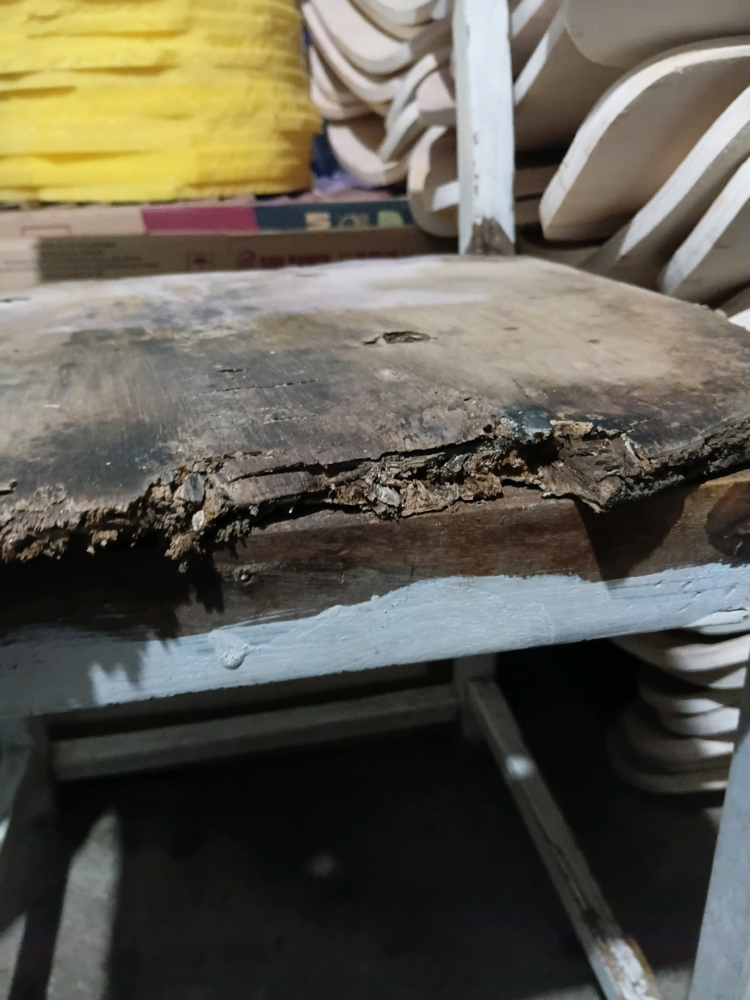 The image shows a close-up of the damaged seat of a wooden chair. The wood is weathered and shows significant signs of rot and splintering along the edge. The chair appears to be old and possibly discarded or awaiting repair. In the background, stacked wooden pieces and a roll of yellow material are visible, suggesting a workshop or storage area. The scene is dimly lit, casting shadows that obscure further details of the environment. The image's content does not provide any information regarding its location in Cikal, Indonesia.