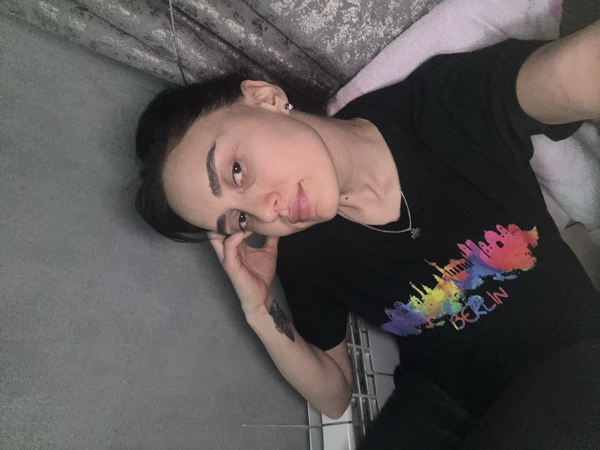 A woman is lying down. Her head is resting on her hand. She is wearing a black t-shirt with a colorful Berlin skyline design on the front. She has dark hair and is wearing earrings and a necklace. Her arm has a tattoo on it. She is lying on a gray surface with a pink pillow behind her. The location is not identifiable as being in Tbilisi, Georgia.