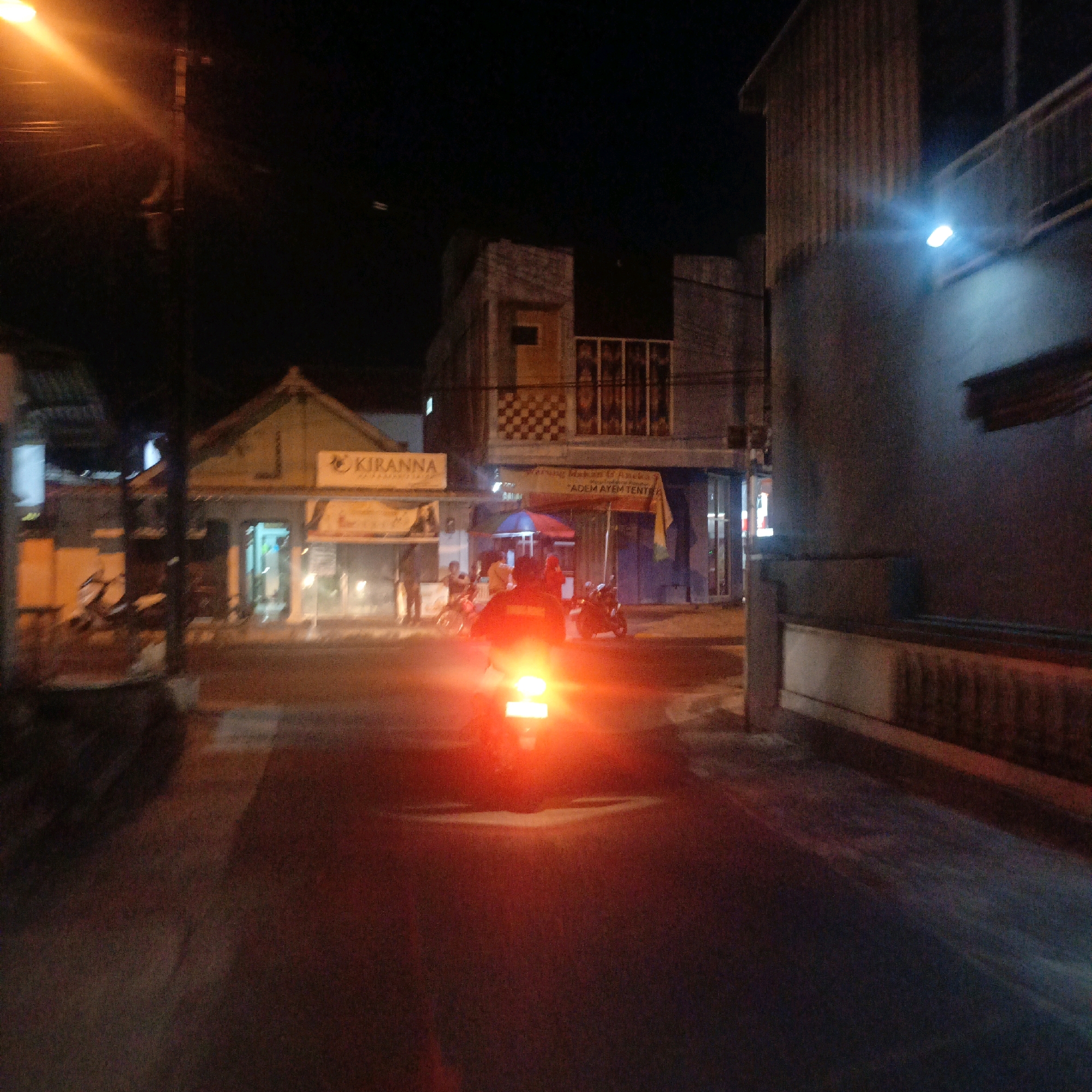 The image depicts a nighttime street scene in Salatiga, Indonesia, with a focus on a person riding a motorcycle. The motorcycle's taillight emits a bright, intense red glow that dominates the foreground and creates a strong lens flare. The rider is silhouetted against this light, making their features indistinguishable. Several other motorcycles are visible further down the street, some with riders.
To the left of the street, there is a building with a sign that reads "KIRANNA". This building appears to be a commercial establishment, possibly a shop or restaurant, with illuminated windows. Adjacent to it, there is another building with a red awning and a sign in Indonesian that reads "ADEM AYEM TENTREM". Further in the background, other buildings are visible, some with visible architectural details and lights.
The street itself is paved and appears wet, reflecting the ambient light and the bright taillight of the motorcycle. The overall atmosphere is that of a typical urban night scene, with artificial lighting from streetlights and buildings illuminating the surroundings. A lamppost with an orange light source is visible on the left side of the frame, casting a beam of light. Another streetlight, emitting a bluish light, is visible on a building to the right. The image is somewhat blurry, suggesting it may have been taken while in motion.
