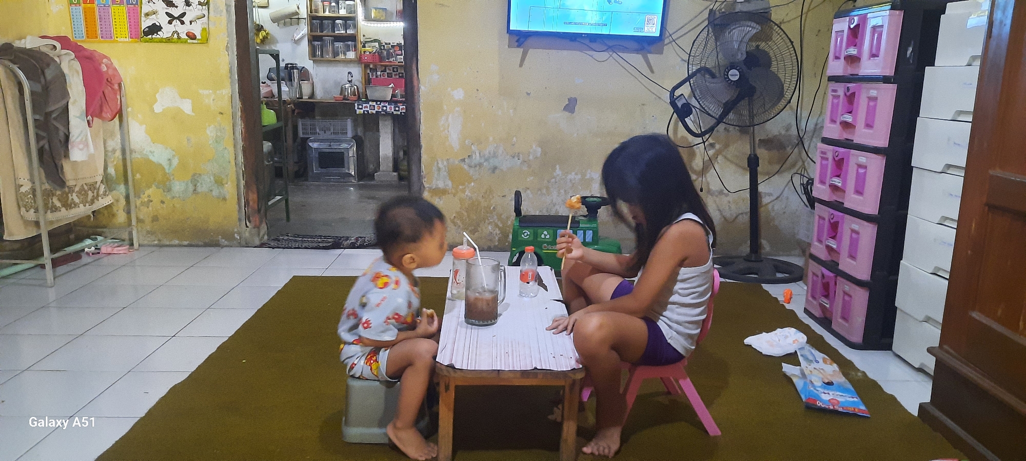 In Ploso, Indonesia, a young boy sits on a grey stool to the left of a low wooden table, facing right. He wears a light blue shirt with cartoon characters and matching shorts. On the table, there's a glass with a straw containing a brown beverage, a bottle with a pink label, and a small carton. To the right of the table, a girl sits on a pink chair, facing the table. She wears a white tank top and purple shorts. She holds a skewer with a piece of food on it. Behind the table and chairs, a green toy vehicle is visible. A standing fan is positioned behind the girl. The room has yellow walls with peeling paint and a dark brown carpet covers the floor. To the far left, a clothing rack holds several garments. A television is mounted on the wall above the fan. To the right, a pink and black plastic storage unit with drawers stands next to a white storage unit.