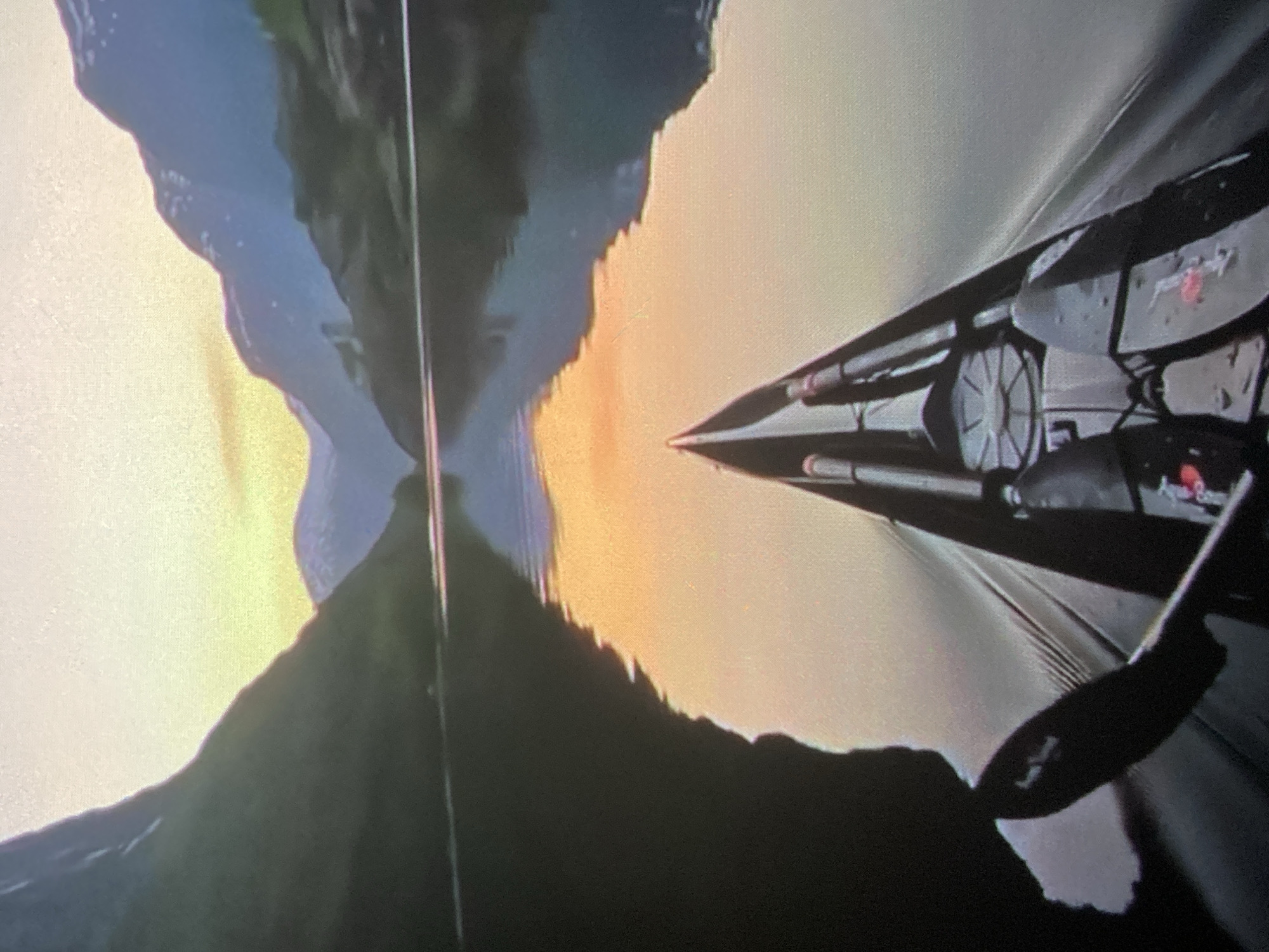 This image shows a first-person perspective of someone in a kayak on a calm body of water, possibly a lake or a fjord. The kayak is dark-colored with paddles resting within it. The water reflects the sky and the surrounding mountains, which are dark green and rocky, with some snow-capped peaks in the distance. The sky appears to be during sunrise or sunset, with warm orange and yellow hues reflecting on the water. The overall scene is serene and natural, with no signs of urban development or people other than the presumed kayaker. The location is not identifiable as Ustyvytsia, Ukraine, as there are no specific landmarks or features to confirm this.