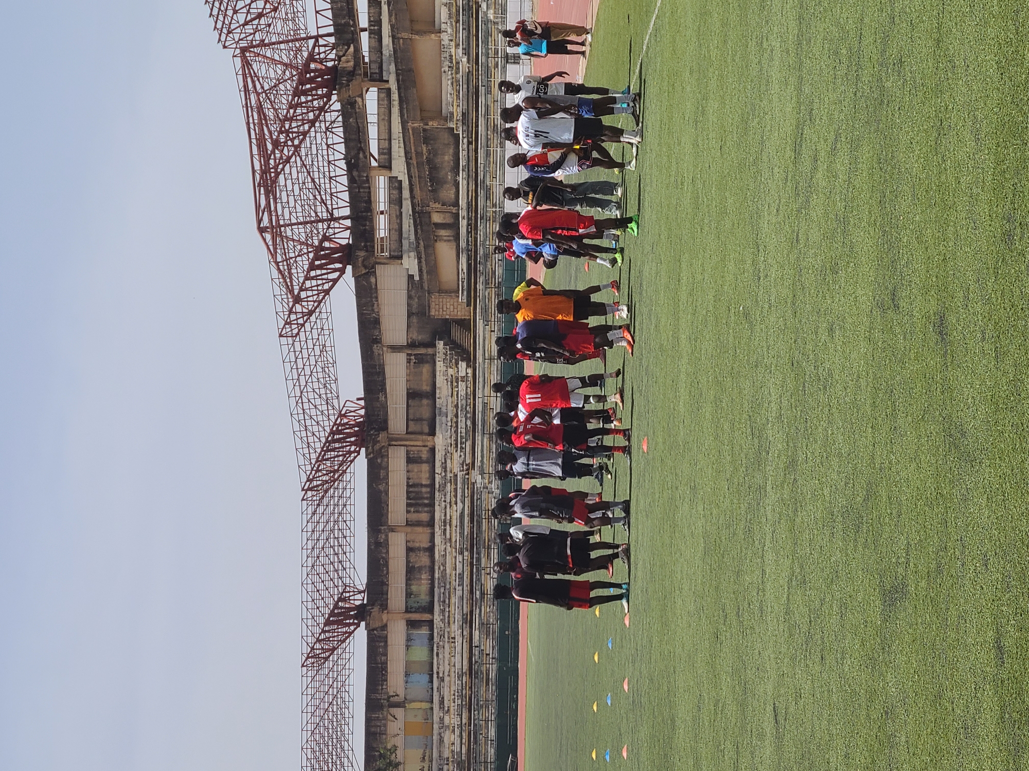 The image shows a group of soccer players lined up on a green soccer field next to the New Stadium in Nigeria. The stadium's weathered concrete structure with red metal scaffolding rises beside the field. The players are wearing various colored jerseys and shorts. Multi-colored cones are scattered on the grass in front of them. The sky is pale blue.