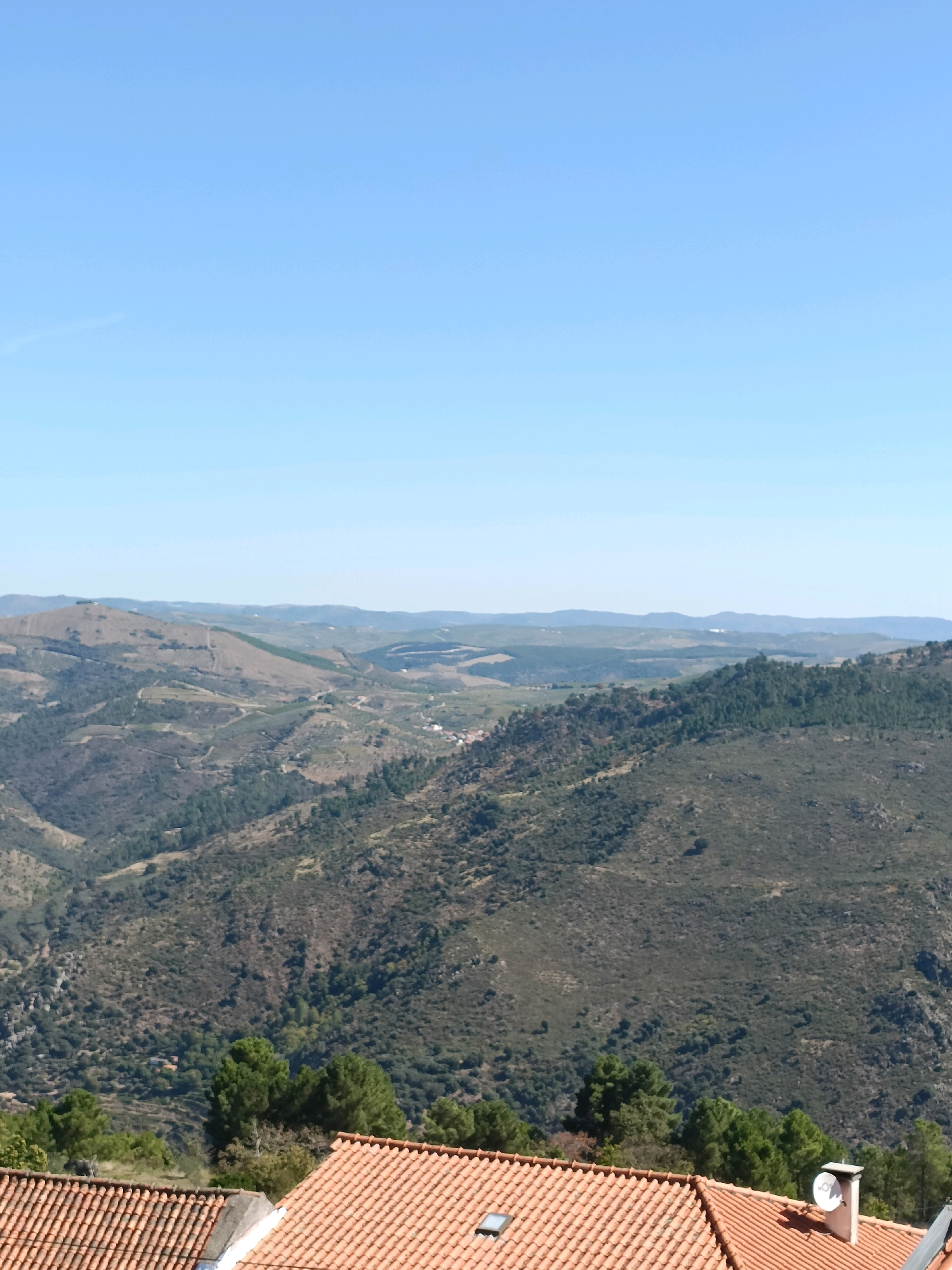 The image shows a panoramic view of a valley landscape with rolling hills and mountains under a clear blue sky. In the foreground, the terracotta tiled roofs of several buildings are visible. A white chimney with a satellite dish attached to it stands on one of the roofs. The middle ground is dominated by the lush green slopes of the valley, densely covered with trees. The background consists of further undulating hills fading into the distance. The scene appears to be set in a rural area, likely in or near Tabuaço, Portugal, given the architectural style of the roofs and the Mediterranean landscape. There are no people or animals visible in the image. The overall impression is one of serene natural beauty and rural tranquility.
