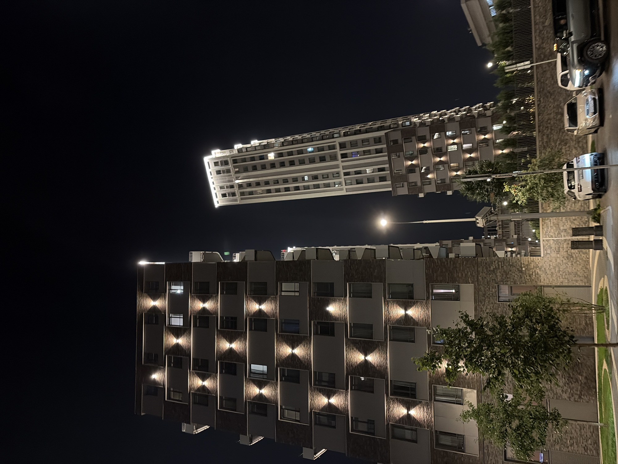 In Moscow, Russia, at night, a low-angle, wide shot captures two modern high-rise apartment buildings. The building on the left is shorter and wider, featuring a facade with alternating bands of grey panels and brown brickwork, illuminated by strategically placed exterior lights that create dramatic shadows. The building on the right is a much taller, slender skyscraper with a white and grey facade, its top illuminated by a bright white light, and its many windows reflecting light from within. In the foreground, several cars are parked on a paved area in front of a low stone wall, with a street lamp casting a warm glow on the scene. Trees with full green foliage are visible, adding a touch of nature to the urban landscape. The sky is a deep, dark blue, indicating late evening or night.