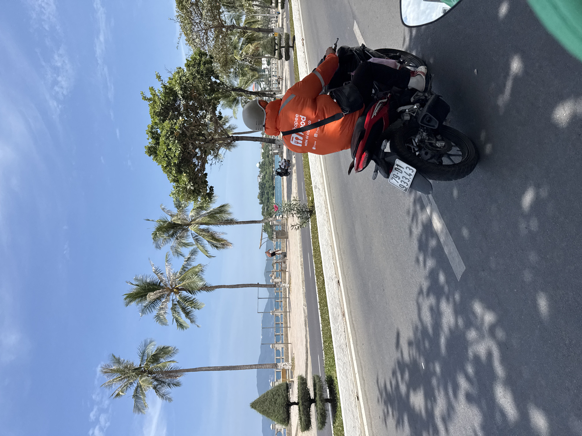 This image shows a street scene in Nha Trang, Vietnam, likely along the coast. In the foreground, a person wearing an orange Shopee Food jacket and a grey helmet is riding a red and black motorcycle. The motorcycle has a license plate that reads "79-D1 933.43". The rider is looking forward, and a black bag is slung over their shoulder. The road is asphalt, with some shadows from trees cast upon it.
In the background, a pedestrian walkway is visible, lined with palm trees and other vegetation. Beyond the walkway is the sea, with a clear blue sky overhead. Hills can be seen in the distance. Several other people and motorcycles are also present in the background, suggesting a populated area. The overall environment appears to be a bright, sunny day.