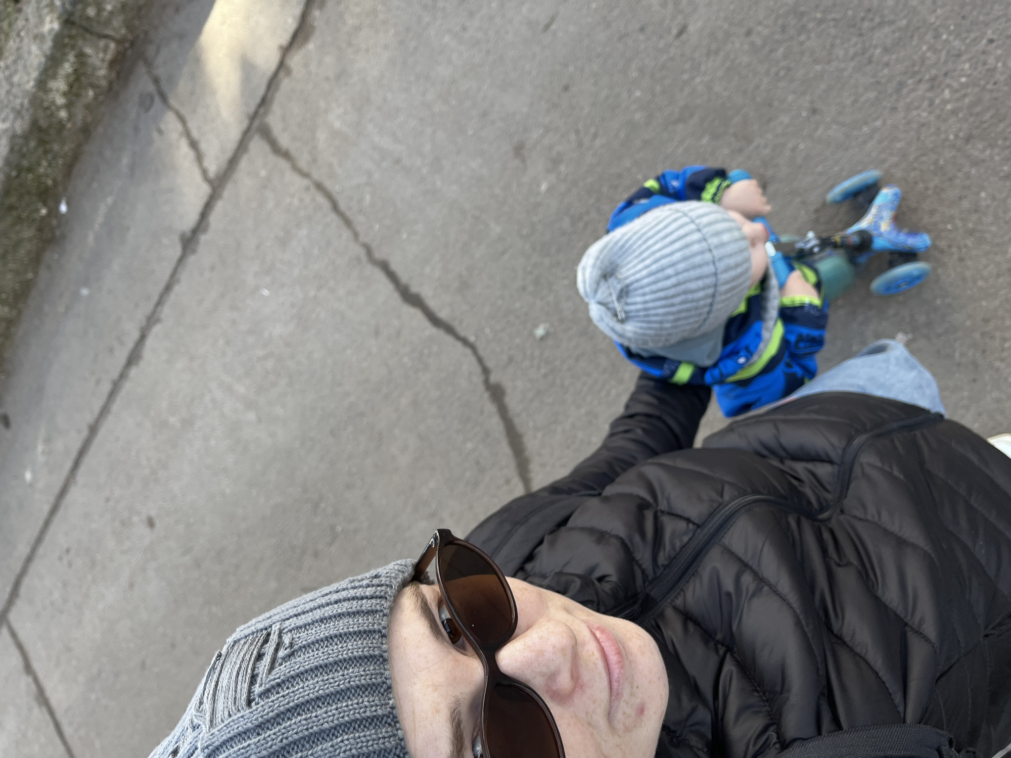 A high-angle shot captures two individuals on a cracked concrete pavement, potentially in an urban setting. The foreground features a person wearing sunglasses and a grey knitted hat, with a black puffer jacket visible. The other individual, likely a child, is further away, wearing a blue and green jacket and a similar grey knitted hat, riding a three-wheeled blue scooter. The concrete is cracked with visible curb to the left. It's not possible to confirm the location as Chaikovsky, Russia, from the image alone.