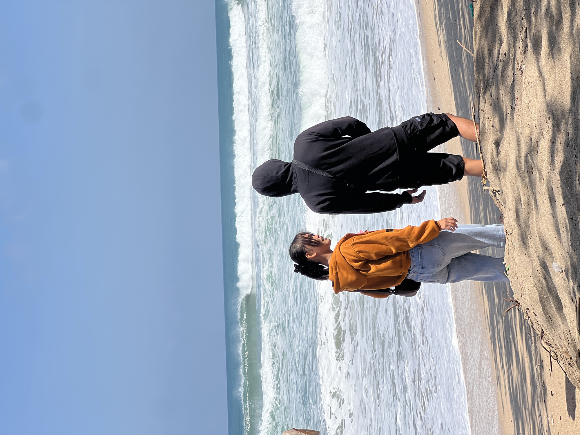 Two individuals stand on a sandy beach, facing the ocean. A person on the left, with dark hair pulled back in a ponytail, wears an orange hooded sweatshirt and light blue jeans. They are looking to their right, towards the other individual. The person on the right, dressed in a black hooded sweatshirt and black shorts, faces away from the camera, observing the waves. The ocean exhibits distinct lines of waves breaking and white foam. The sky is clear and blue. The beach in the foreground is composed of sand, with scattered dry vegetation and shadows cast by unseen trees. The overall scene depicts a coastal environment with people enjoying the view of the sea.