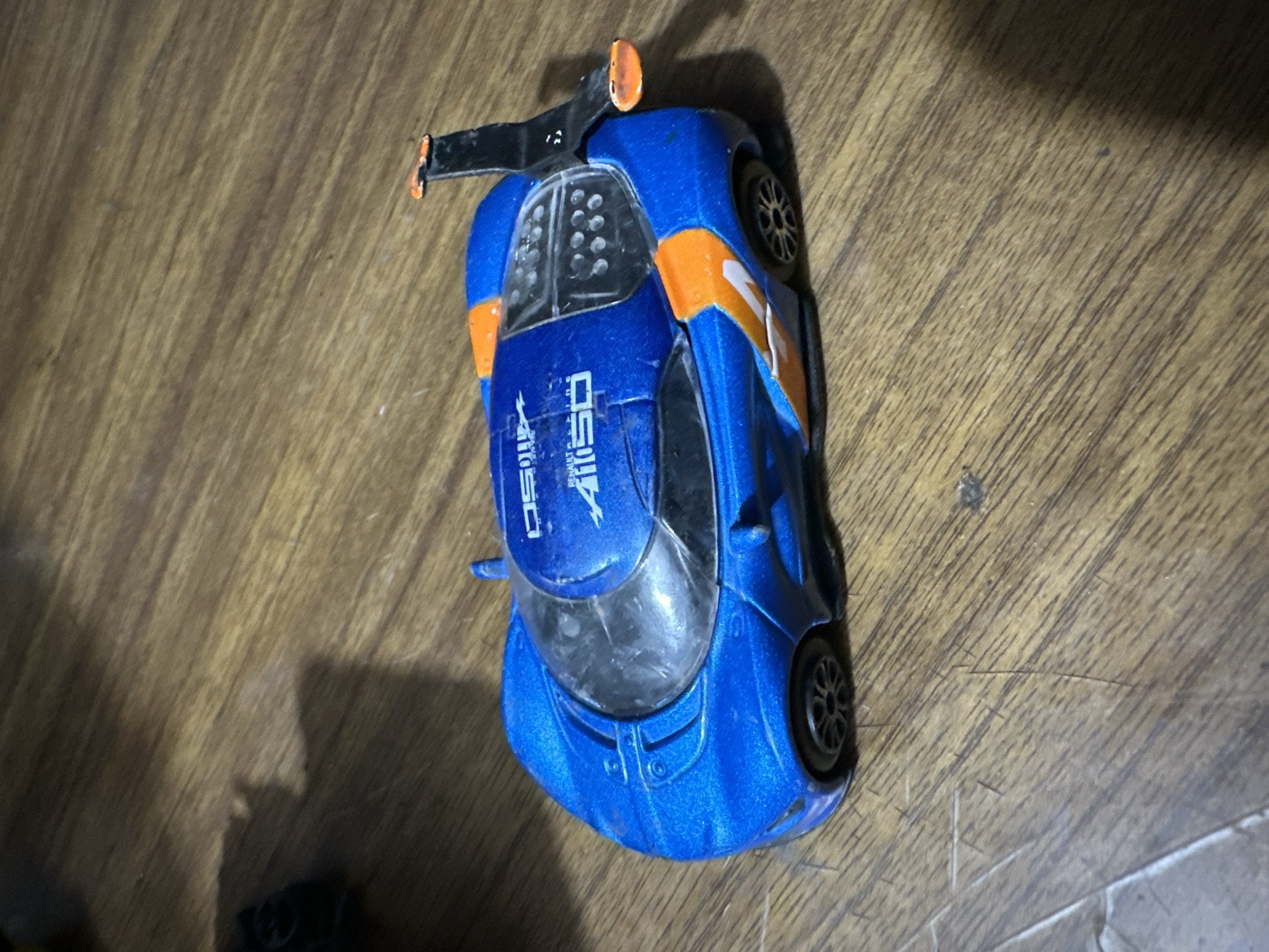 A blue toy race car with orange accents is positioned on a wooden surface. The car has a prominent spoiler on its rear, with an orange tip. The car's body is adorned with white decals. One decal features a stylized graphic and the number "1150", with smaller text that appears to read "BEHANCE STUDIO". Another decal on the side shows a white "1". The wheels are black with silver rims. The wooden surface has a visible grain pattern and appears to be a tabletop or floor. The lighting suggests an indoor setting, possibly with artificial light casting shadows. There are no people or animals visible in the image. The overall scene is a static, still life arrangement of the toy car.