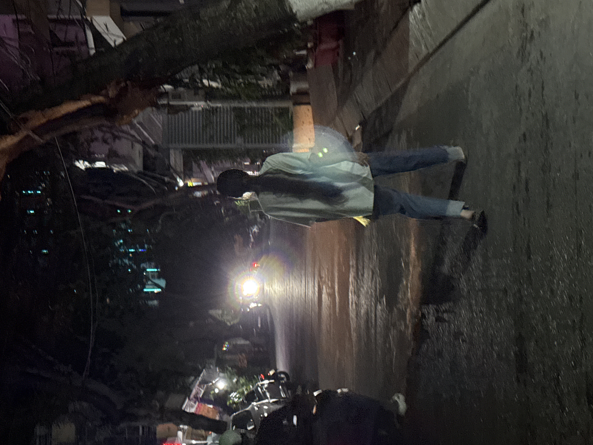 In the city of Bac Nha Trang Ward, Vietnam, a person with long, dark hair is walking away from the camera at night on a street. The person is wearing a light-colored, long-sleeved shirt and blue jeans that are rolled up at the cuffs. They are also wearing dark shoes. In the background, a bright light from a vehicle is visible, illuminating the street. Trees line the side of the street, and there are buildings and other vehicles in the distance. The ground is wet, reflecting the lights.