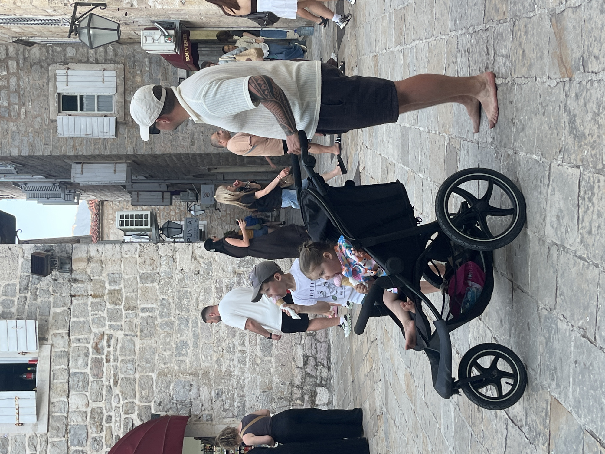 The image depicts a street scene in Budva, Montenegro. In the foreground, a man wearing a white cap and a light-colored polo shirt with dark shorts stands barefoot, looking to the right. He is pushing a black stroller. Inside the stroller is a young child, a girl wearing a colorful outfit, holding an ice cream cone. A boy, also holding an ice cream cone, stands next to the stroller. The background shows a stone building with windows and shutters, and other people walking in the street. The ground is paved with cobblestones.