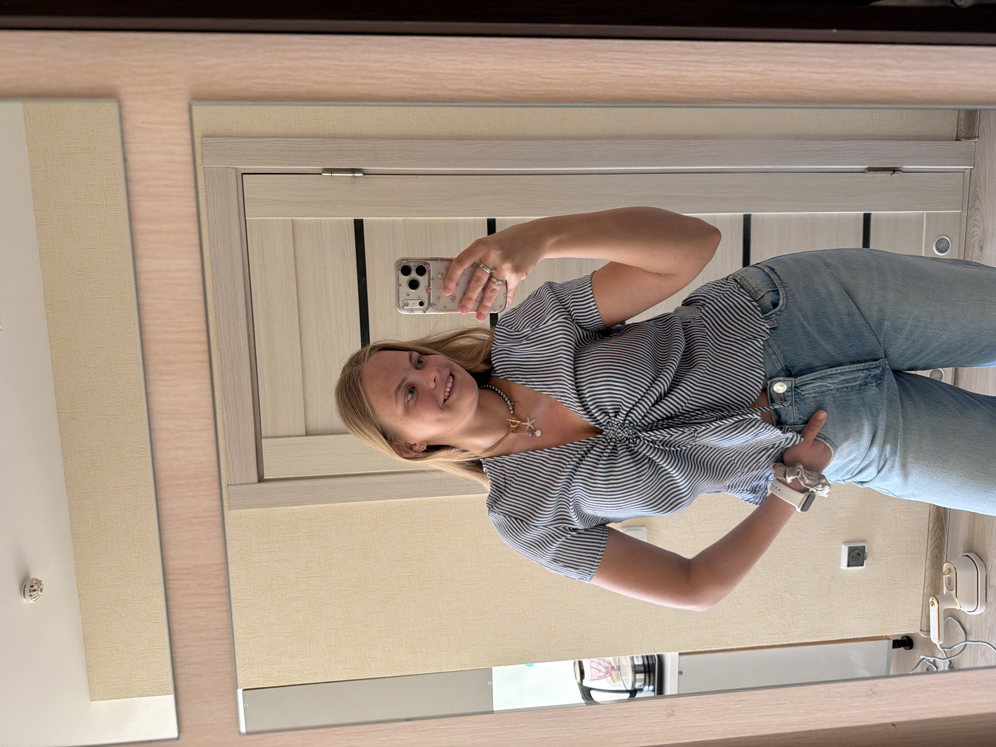 A woman is captured in a mirror selfie within what appears to be a residential interior in Izhevsk, Russia. She is smiling broadly and looking directly at the camera phone she holds with her right hand. Her blonde or light brown hair is parted on the side. She is wearing a short-sleeved, blue and white striped top that is cinched at the front, light-wash blue jeans, and a beaded necklace with a gold-colored charm (possibly a star or anchor). On her left wrist, she wears a white smartwatch and a light-colored scrunchie. Several rings are visible on her right hand.
The mirror reflects a room with light beige or cream-colored textured wallpaper on the walls. Behind the woman, a light-colored wooden door with vertical panels and matching trim is visible. A white electrical outlet is on the wall behind her hip, and to its right, a small white and gold appliance, possibly a mini steamer or iron, is plugged in. In the upper left corner of the reflected image, a white smoke detector is visible on the ceiling. The overall lighting is bright, suggesting daytime or a well-lit indoor environment. No explicit text is clearly visible in the image. The woman's pleasant expression suggests a positive mood.