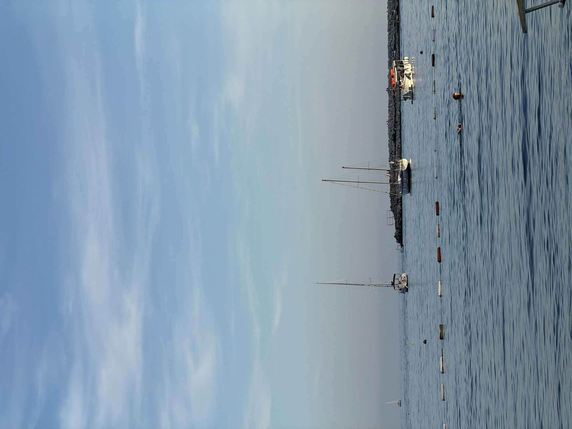 The image displays a serene coastal scene in Budva, Montenegro, under clear daytime conditions. The view is primarily composed of a calm body of water, likely the sea, in the lower half of the frame, and a clear blue sky above.
The water's surface is gently rippled, reflecting the bright sky. Numerous buoys, mostly white and orange, are scattered across the water, suggesting a marked area for swimming or navigation. Two individuals are visible swimming in the water, their heads and upper torsos breaking the surface.
A dark, rocky breakwater or natural shoreline stretches horizontally across the midground, separating the open water from the backdrop. Moored near this shore are several boats. Two prominent sailboats, one white and one darker, are anchored with their tall masts reaching skyward. Closer to the rocky edge, a white motorboat is visible, adorned with several bright orange life vests. Other smaller vessels are also discernible along the shoreline.
The upper portion of the image features a expansive light blue sky, with delicate, wispy white clouds adding texture. The bright and even lighting indicates it is daytime, likely mid-morning or early afternoon, under fair and pleasant weather. The overall atmosphere is one of tranquility and leisure, typical of a Mediterranean resort location. There is no visible text within the image.