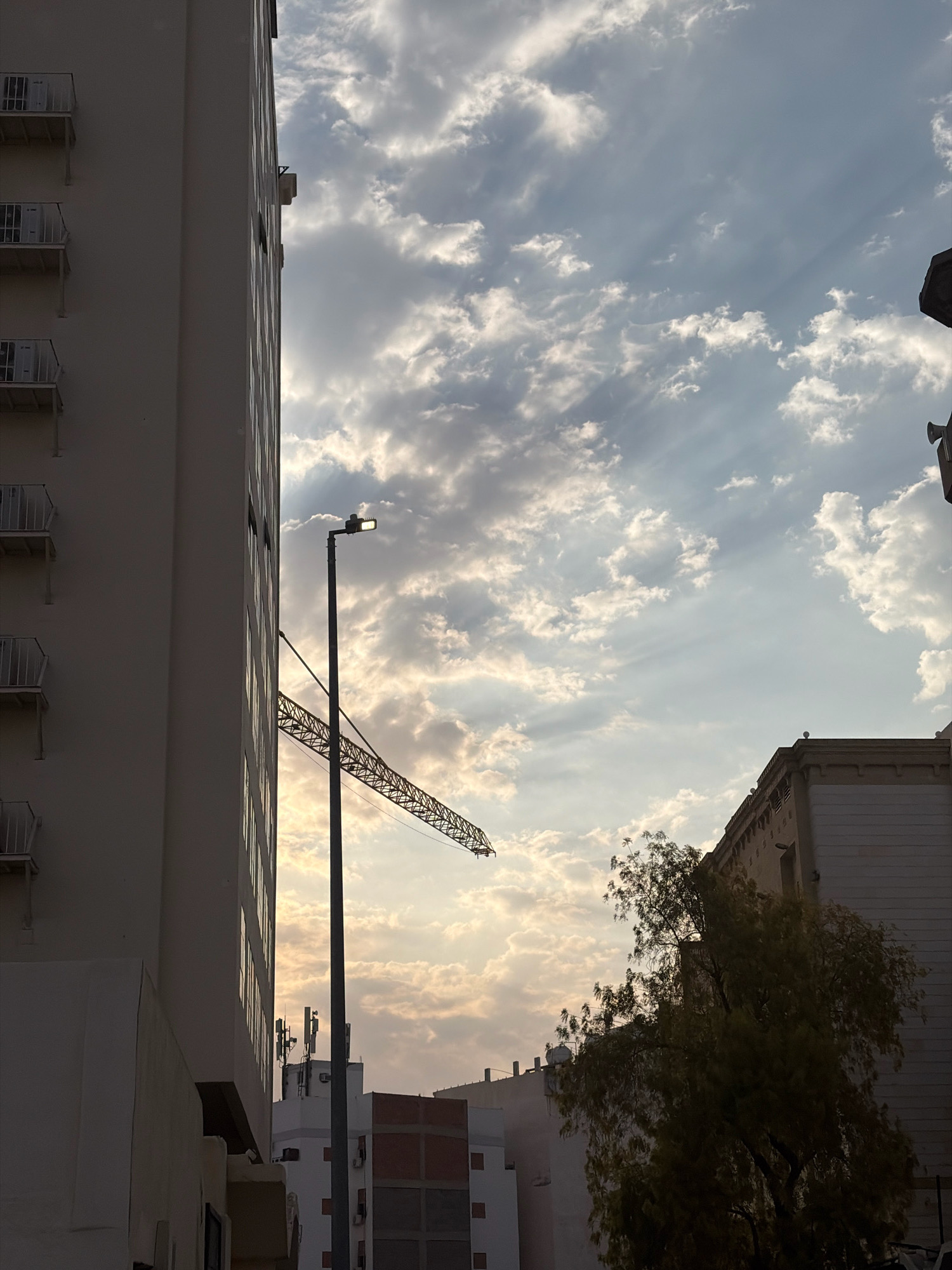 In the city of Medina, Saudi Arabia, a tall, pale building dominates the left side of the frame, its facade punctuated by rows of balconies with white railings. A streetlamp stands in front of the building, its light illuminated, and behind it, the arm of a construction crane extends diagonally across the scene. The sky above is filled with dynamic clouds, some wispy and others more substantial, with rays of sunlight breaking through, creating a dramatic effect. In the lower right corner, a portion of another building is visible, along with a dark, leafy tree. The overall impression is one of urban construction and natural beauty coexisting.