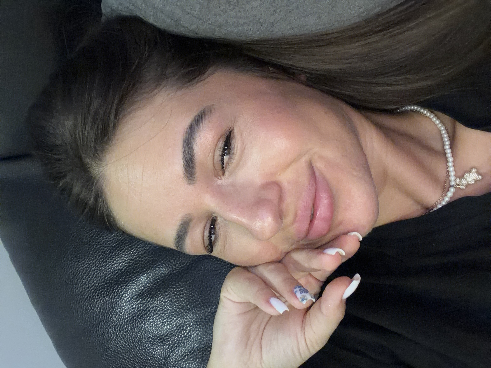 The image is a close-up, high-angle, indoor shot of a woman's face and upper body. She has brown hair, fair skin with noticeable makeup, and is wearing a black top and a pearl necklace with a teddy bear pendant. Her left hand is resting on her cheek, and her fingernails are painted white with a floral accent on one nail. The background appears to be a black leather couch on the left and a gray surface on the right, possibly a pillow or another piece of furniture. There is no visible indication of the city of Novi Iskar, Bulgaria, or any specific urban or outdoor environment.
