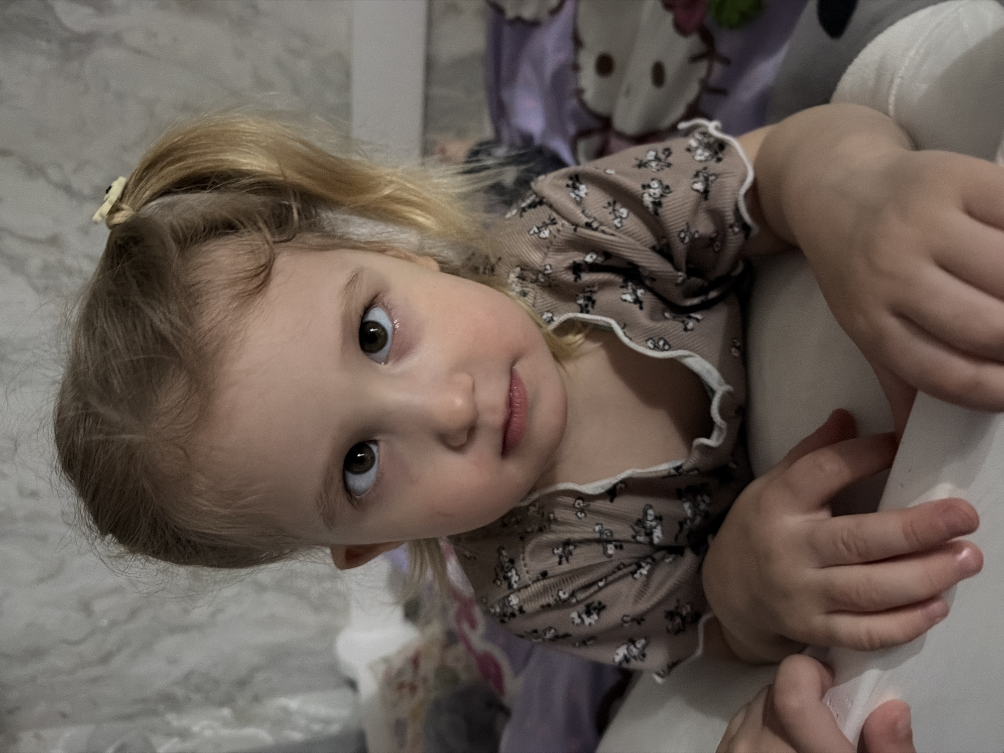 A young child with blonde hair pulled into a ponytail and secured with a small, light-colored clip is looking directly at the camera. The child is wearing a beige long-sleeve shirt with a small floral print and white ruffled trim around the neckline. The child's hands are resting on a white, soft object. In the background, there is a purple fabric with an image of Hello Kitty and a gray, marbled surface. The setting and context of the city of Noviy, Russia, cannot be determined.