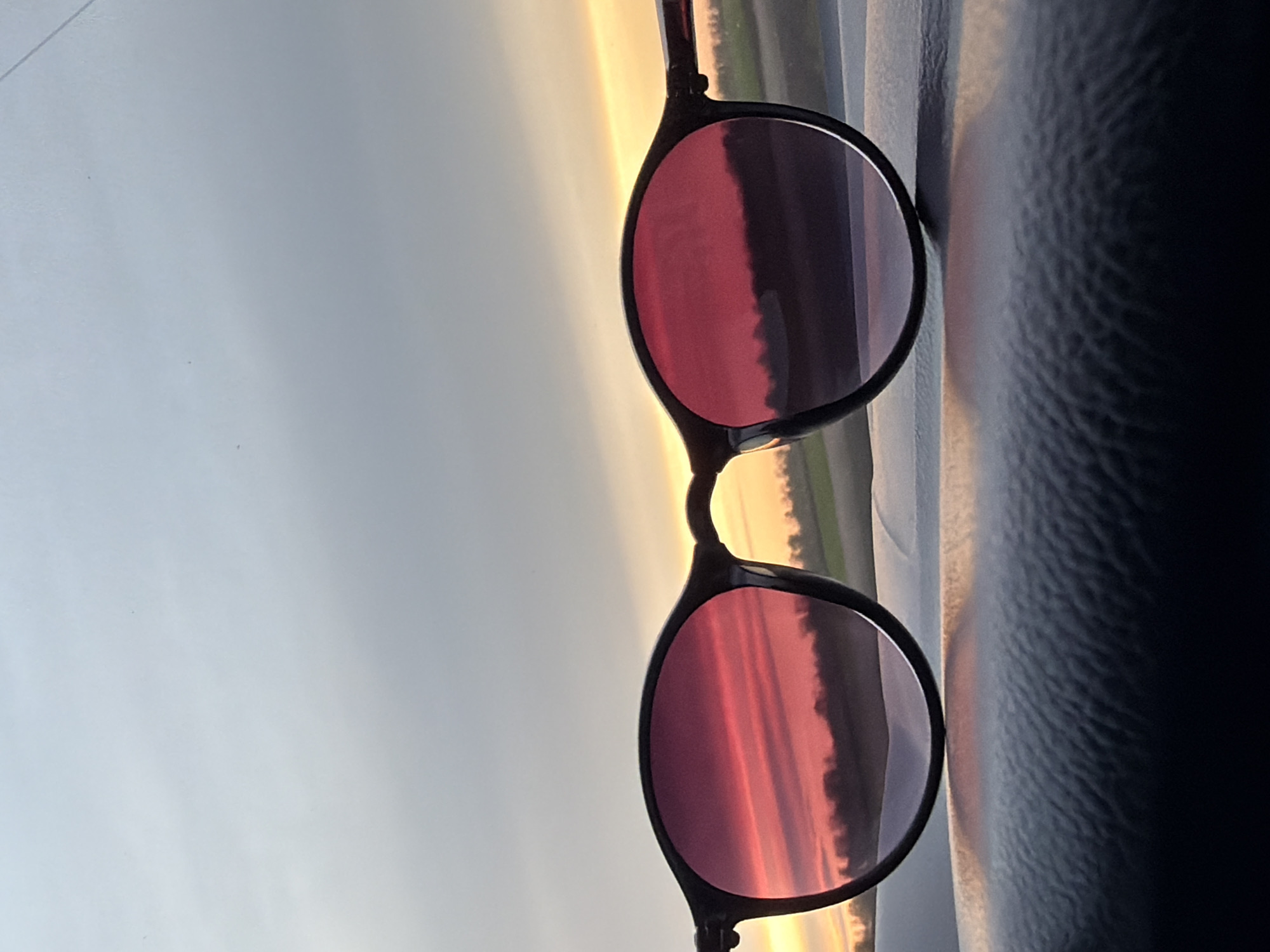 A pair of black-framed sunglasses is captured, appearing rotated 90 degrees clockwise from a natural horizontal orientation. The lenses vividly reflect a colorful sky at either sunrise or sunset, dominated by hues of reddish-pink, orange, and purple. Within these reflections, a dark, undulating landscape, possibly fields or distant trees, is silhouetted along the horizon.
Beyond the sunglasses, the actual sky displays a softer gradient of light blue, grey, and golden-yellow light towards the horizon. To the right, a dark, textured surface, resembling a car interior such as a dashboard or seat, is visible, suggesting the photo was taken from inside a vehicle looking out. The scene conveys a tranquil moment at dawn or dusk in Alexandrovka, Russia. There is no visible text in the image.