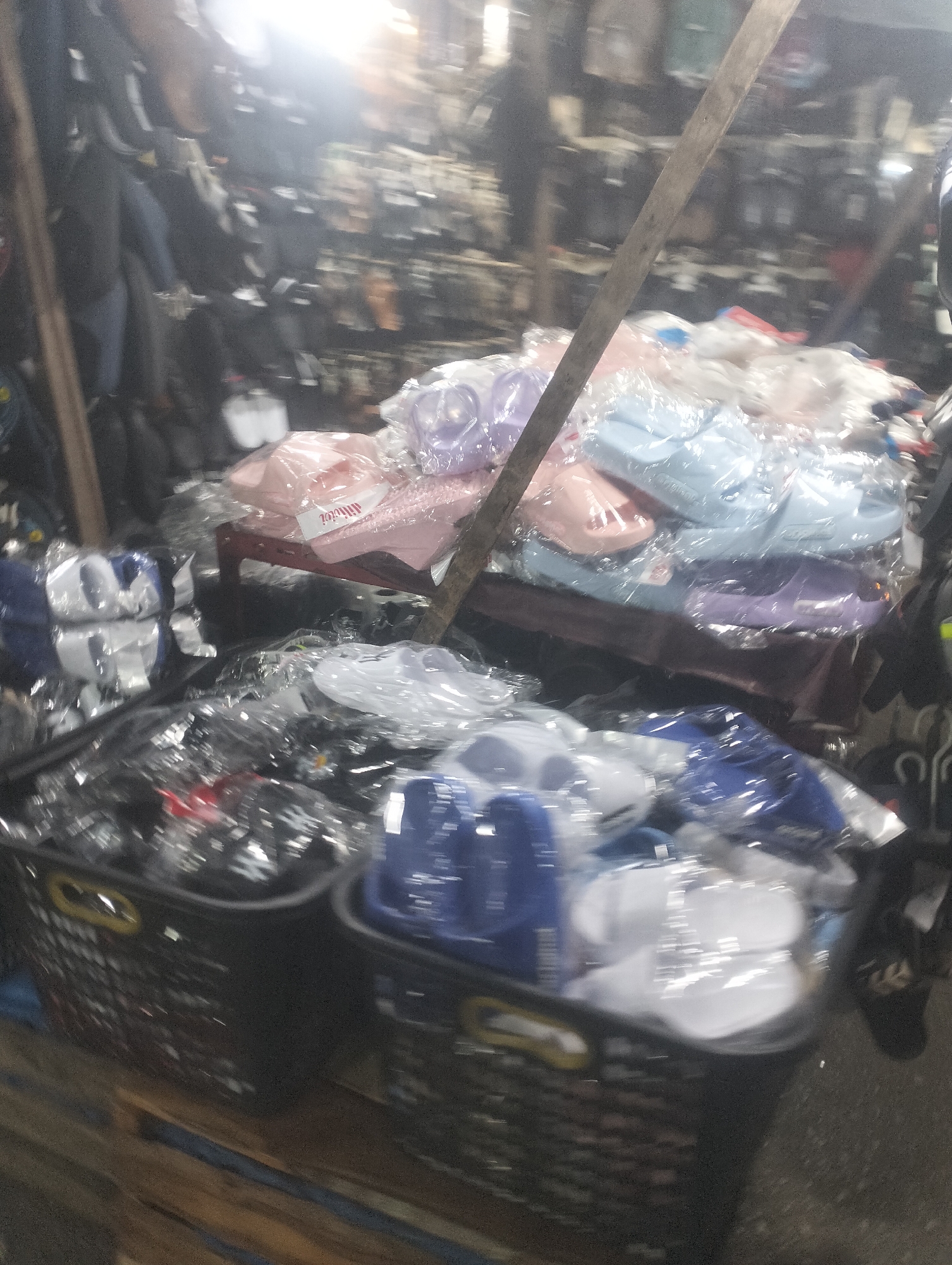 This image depicts a market stall in Batang Serosa, Indonesia, selling various types of footwear. In the foreground, two black plastic baskets are visible, filled with individually packaged flip-flops and sandals. The flip-flops in the left basket are predominantly white and blue, while the right basket contains white and blue flip-flops as well. The packaging is clear plastic, allowing the colors of the footwear to be seen.
Behind the baskets, on a wooden table, a pile of footwear is displayed, also packaged in clear plastic. These include pink, light blue, and purple flip-flops. A wooden pole runs vertically through the scene, obscuring parts of the background.
The background shows a cluttered market environment with shelves filled with more footwear and other items, rendered in soft focus due to motion blur. The lighting is artificial and somewhat bright, creating a glare in the upper part of the image. The overall impression is of a busy, casual retail setting. No people are clearly identifiable in the image.