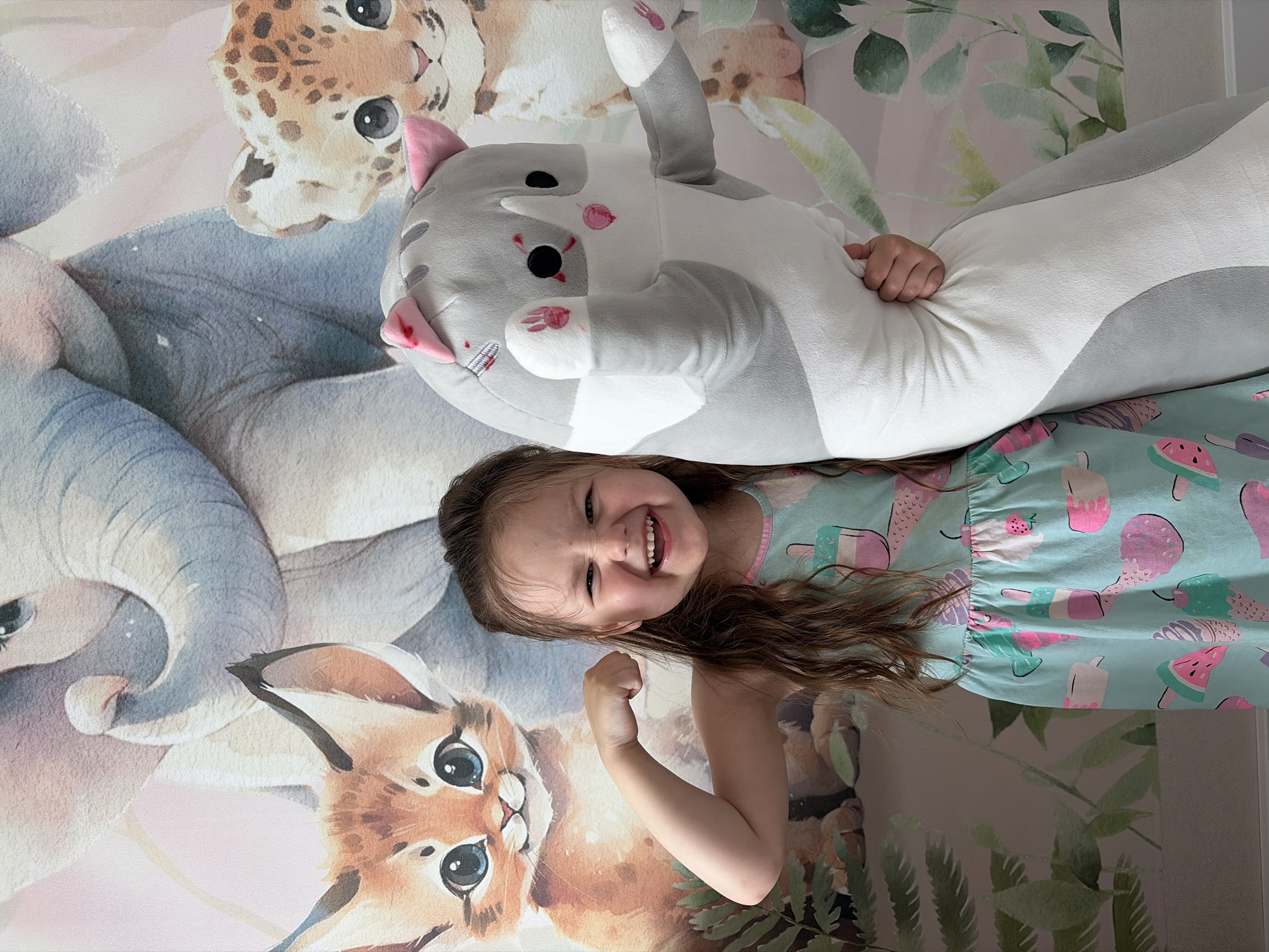 A young girl lies on a patterned surface, possibly a rug or blanket. She is wearing a dress with an ice cream and watermelon design. She is smiling and flexing one arm. A stuffed grey and white cat with pink ears is lying partially on top of her. The background of the surface she is on features drawings of animals including a leopard, elephant, and caracal. Some foliage is also visible in the background. The location cannot be confirmed as Izhevsk, Russia, without further information.