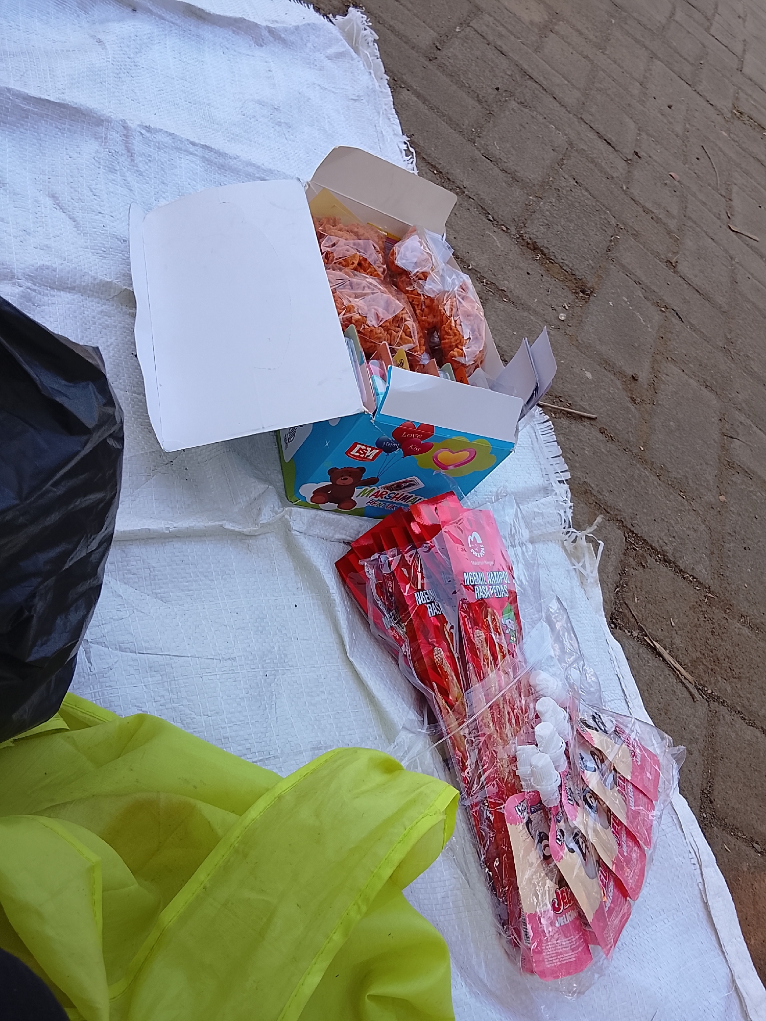 In Krandegan, Indonesia, an outdoor scene shows various food items and materials laid out on a white woven sheet, situated on a paved surface. To the left, a black plastic bag is partially visible, and a bright yellow fabric is spread out. The central focus is an open white cardboard box containing an assortment of orange, puffed snacks in clear plastic packaging. Next to this box is a blue box featuring cartoon drawings of animals and text, likely indicating it holds confectionery or snacks. Below these boxes, several individual pouches of a red product, possibly snacks or drinks, are neatly arranged in clear plastic packaging. These pouches have white caps and are decorated with cartoon imagery. The overall impression is that of a market stall or a display of goods for sale.