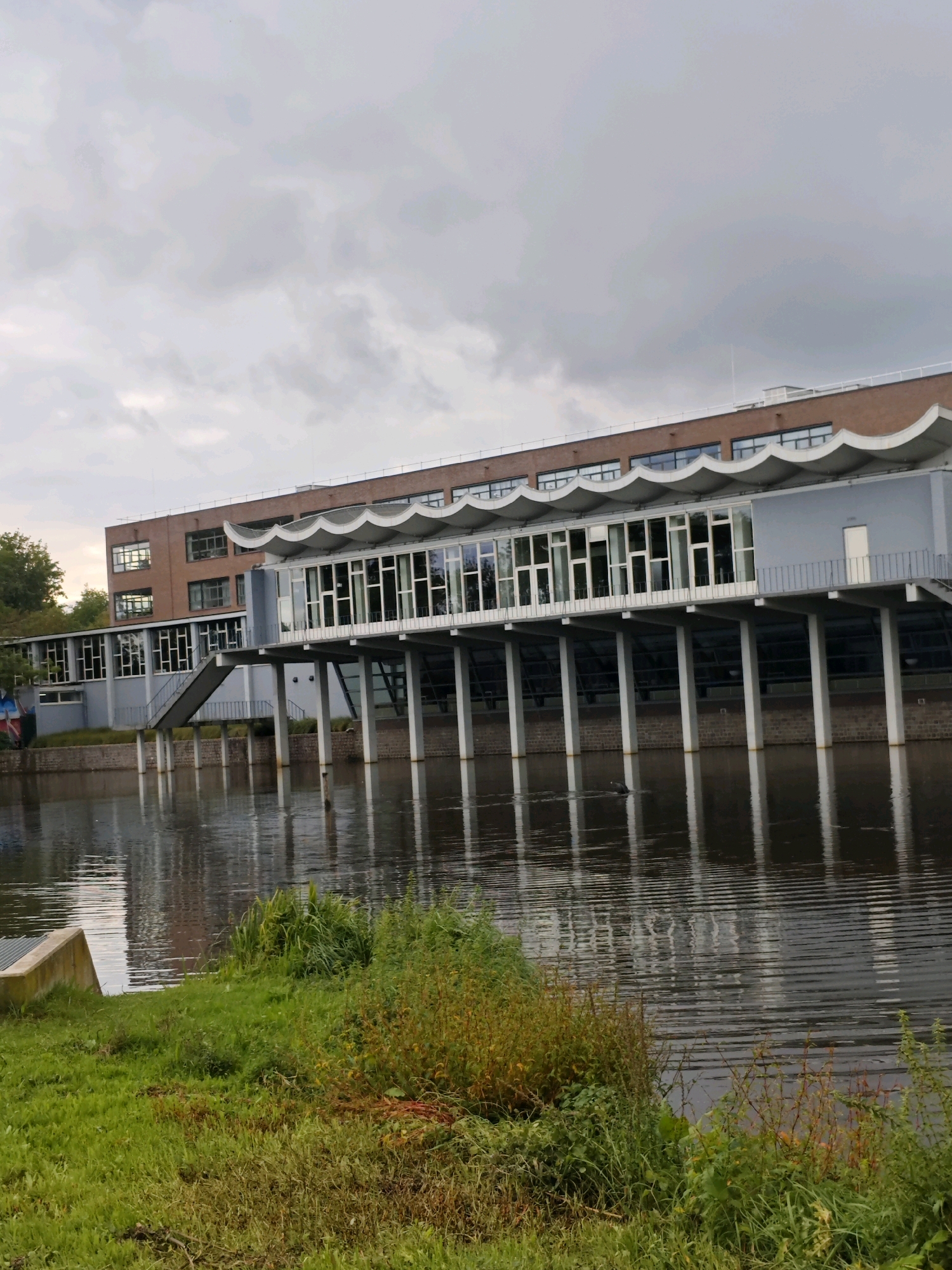 This is a photograph of the former swimming pool building at the Technical University of Eindhoven, located in Eindhoven, Netherlands. The building, designed by Theo van den Broek, features a distinctive wavy roofline and is elevated on stilts over the water. The image captures the building from a low angle, with its facade of large windows reflecting the overcast sky and surrounding water. In the foreground, there is a grassy embankment leading down to the water's edge. The overall scene conveys a sense of architectural interest within an urban setting, with the water acting as a prominent element of the composition.
