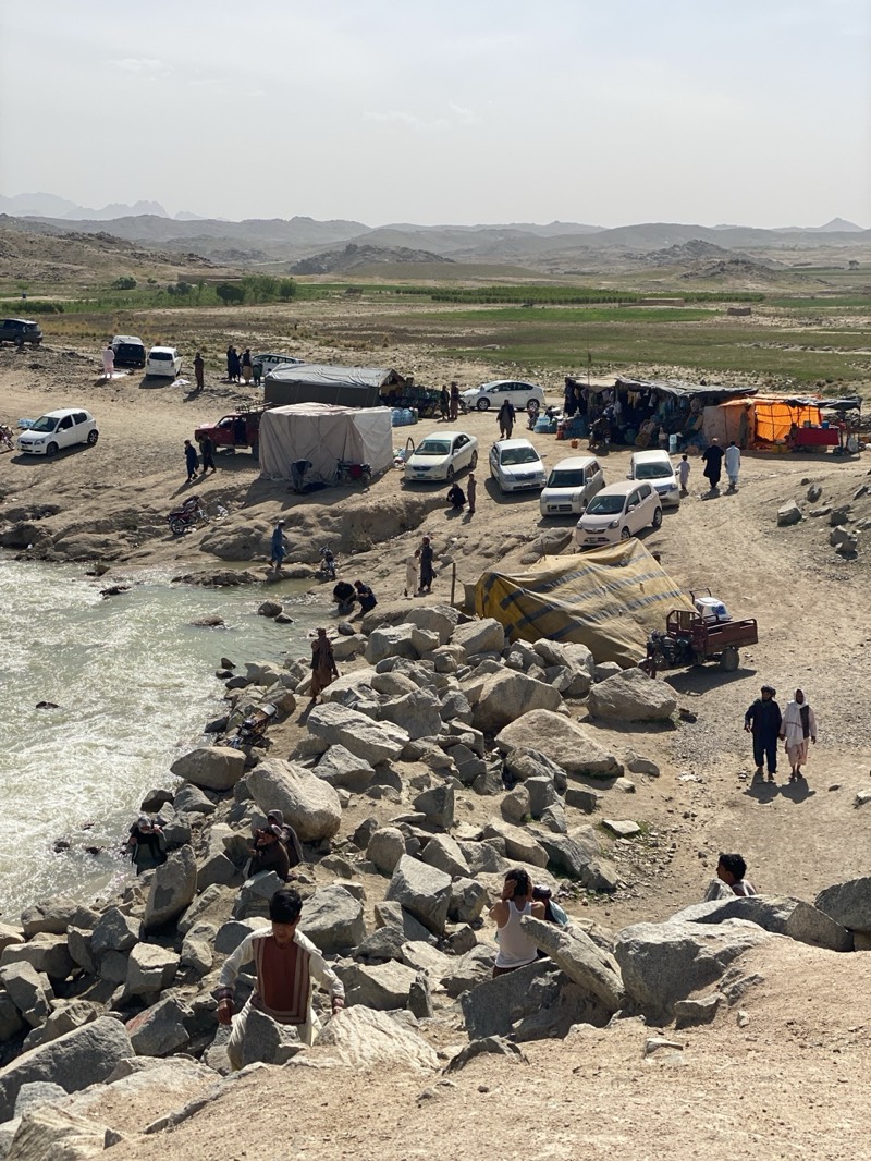 The image shows a scene in a rural area of Kim, Afghanistan. In the background, there are arid hills and a flat green field. Several cars and tents are scattered across a flat, rocky landscape. People are present, some gathered near the tents and cars, others near a river with rocky banks. Some individuals appear to be wading or standing in the water. One person is climbing the large rocks at the bottom of the frame. Other objects include motorcycles, water containers and farm equipment.