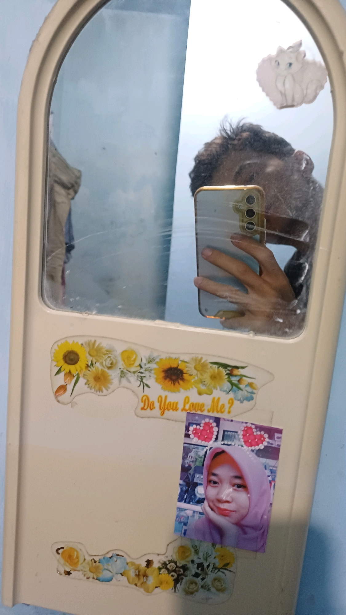 A person is taking a selfie in a mirror in what appears to be a room in Kowangan, Indonesia. The mirror is framed with a beige border and has some scratches. A phone is being held by a hand to capture the reflection of the person's face and the room behind them. On the lower part of the mirror, there are stickers. One sticker is a floral design with sunflowers and the text "Do You Love Me?". Another sticker is a photo of a young woman wearing a pink hijab. The walls in the background are light blue.