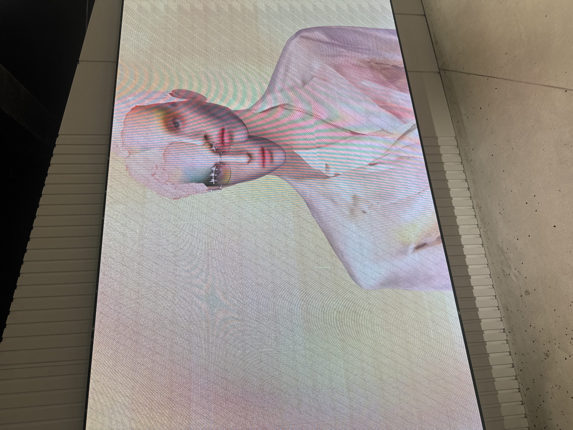 The image shows a large, modern digital screen displaying an abstract digital artwork. The artwork features a distorted, fragmented human face with smooth, mannequin-like skin. The face is rendered in soft pastel colors, primarily pink, white, and pale green, with streaks of color that create a layered, glitchy effect. The subject wears what appears to be a pale pink blazer or jacket. The screen is mounted on a wall with a fluted texture and is positioned above a concrete floor. The scene appears to be an indoor exhibition space or gallery. There is no identifiable information suggesting a specific location in Shanghai, China.