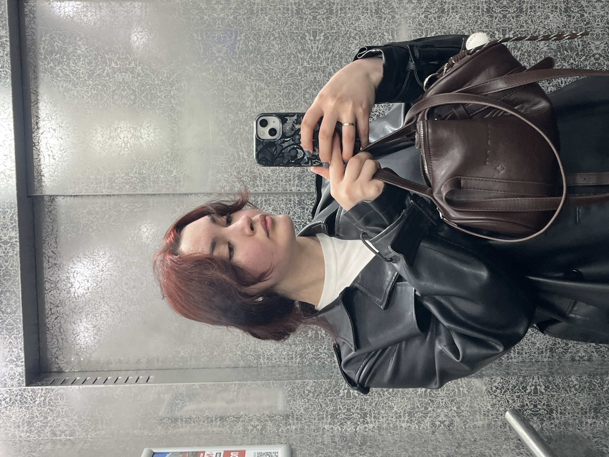 A young woman takes a selfie in what appears to be an elevator with patterned metal walls. She is wearing a black leather jacket over a white shirt, and a brown leather purse is slung over her shoulder. Her hair is short and auburn. The phone in her hand has a patterned black case. An information board is visible at the bottom of the image, with text partially obscured. The scene seems to be indoors, likely in a commercial or residential building. There is no information to pinpoint the location in Neftekamsk, Russia.