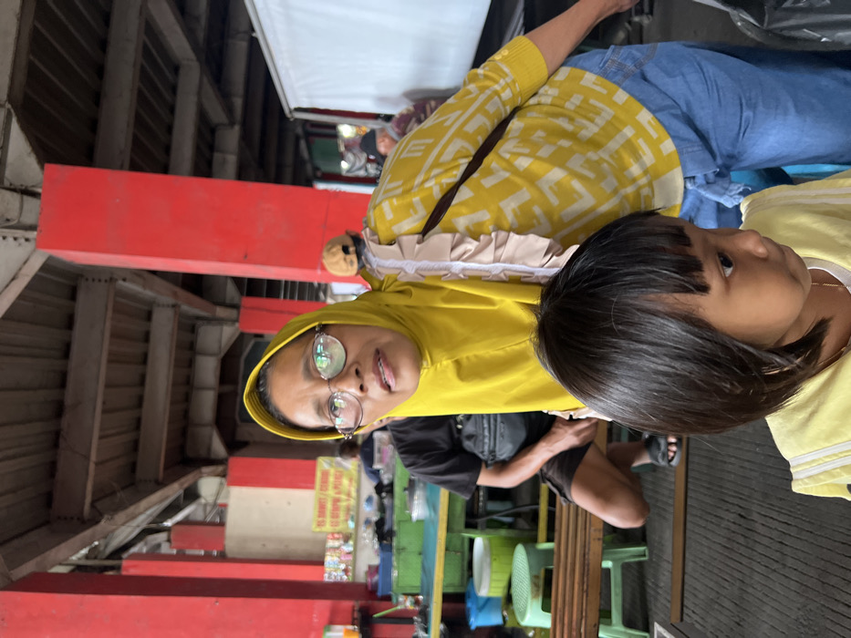 The image depicts a covered market or food stall area in Nganguk, Indonesia. In the foreground, a woman, estimated to be middle-aged, wears a yellow hijab, rimless glasses, a yellow long-sleeved top with a geometric pattern, and blue jeans. A light beige ruffled detail is visible on her right shoulder, and a dark strap crosses her back. Her gaze is directed slightly off-camera to the viewer's right, and her mouth is slightly open. Partially in front of her is a young girl, visible from the shoulders up, with straight dark hair and bangs, wearing a light yellow top. The girl looks downwards and to her right. In the midground, to the left of the woman, a man in a black t-shirt and dark shorts sits on a low wooden bench. The background is characterized by robust red structural pillars supporting an overhead ceiling with visible beams and corrugated material. Behind the main subjects, various market stalls or food preparation areas are discernible, featuring containers, bottles, and a white sign with red lettering, partially readable as "ES DAWET AYU / CENDOL IS KEMBALI MENCE...". Green plastic stools, additional wooden benches, and a large white tarp or barrier are also present in the semi-enclosed commercial setting.