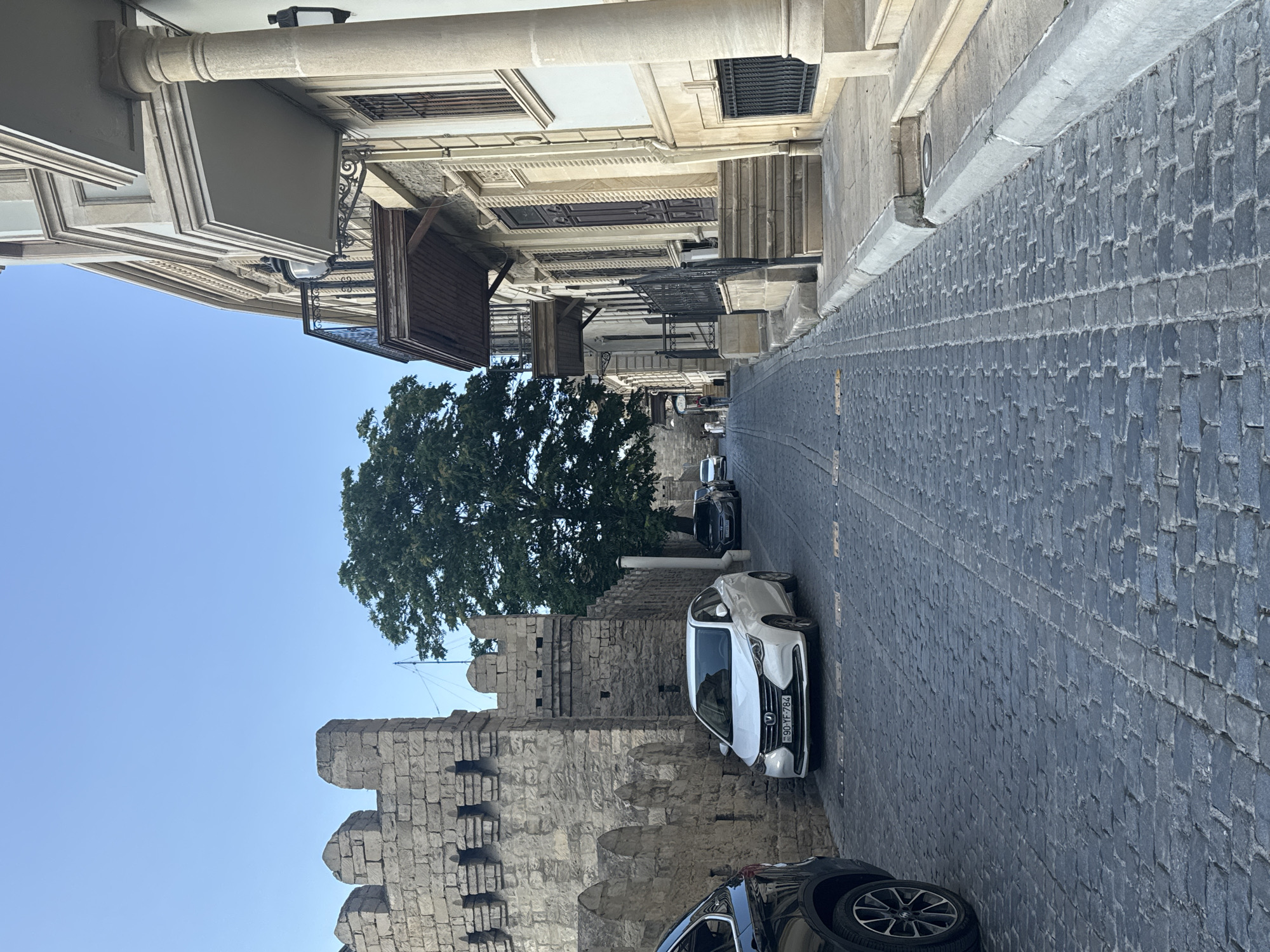 The image shows a narrow, cobblestone street in the city of Sabail Raion, Azerbaijan, likely in the historic Old City (Icherisheher). On the left, a crenellated stone wall, characteristic of ancient fortifications, lines the street. A white sedan is parked in front of the wall, and a black SUV is partially visible on the far left. The right side of the street is dominated by a building with a light-colored facade, featuring multiple stories, balconies with ornate iron railings, and arched windows. A large tree with dense foliage stands between the stone wall and the building. The sky is clear and blue.