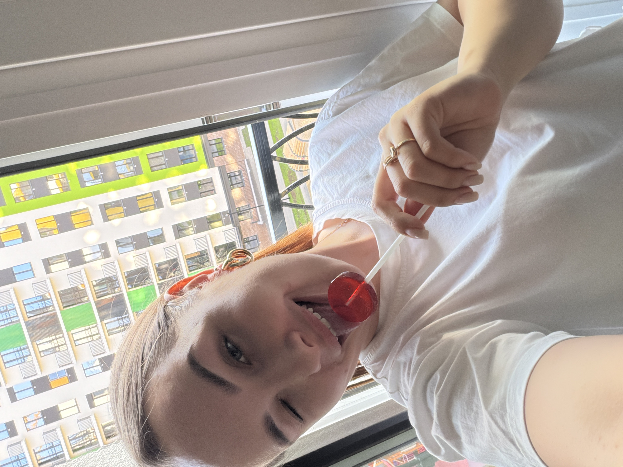 A woman winks while holding a red lollipop to her lips. She is wearing a white t-shirt and a gold ring on her finger. Behind her is a window with a view of a colorful apartment building. The location is Pervomaysky, Russia.