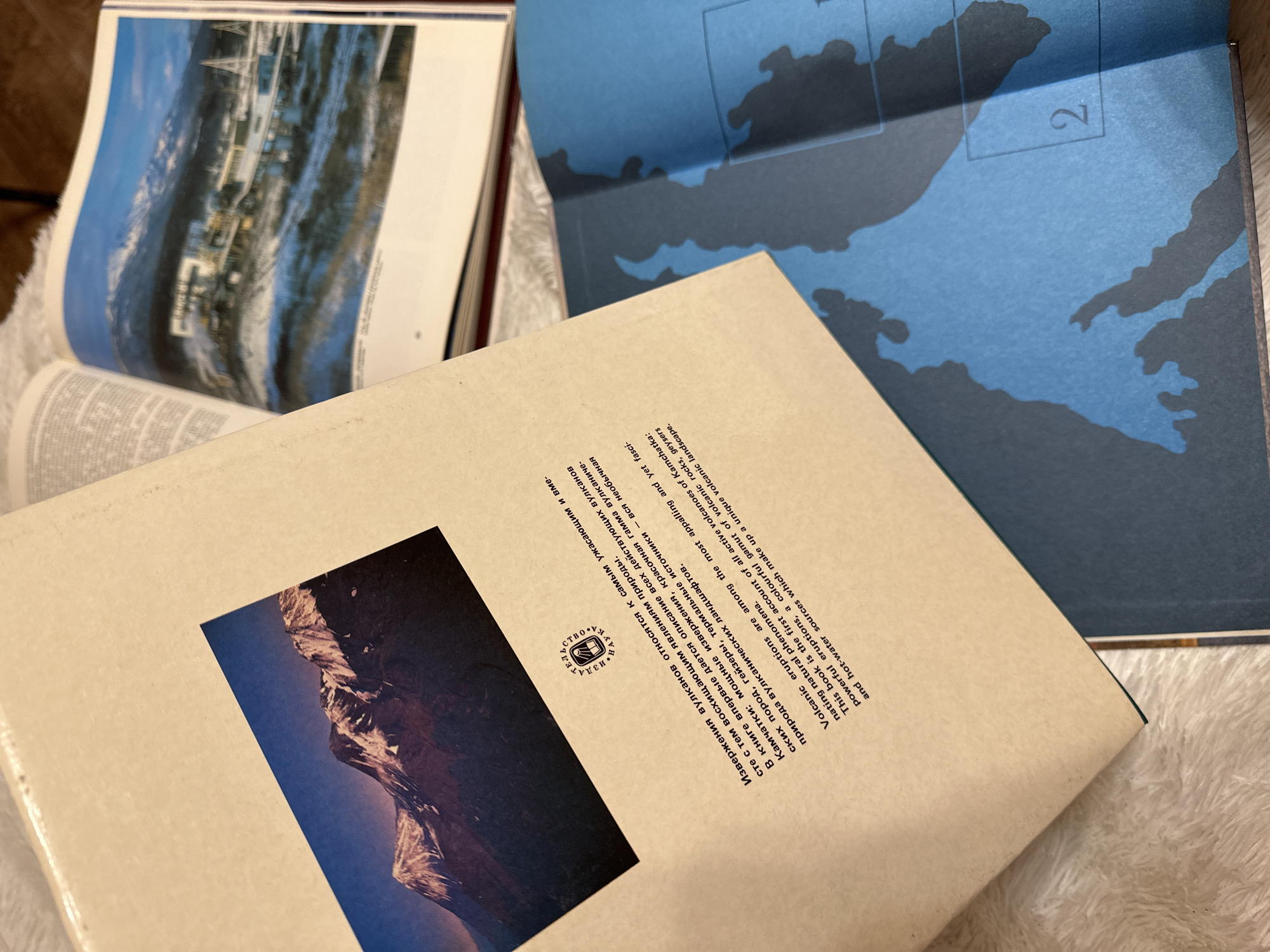 The image shows a close-up of several books. The book in the foreground has a beige cover with a photograph of a mountain range in the bottom left corner and text in what appears to be Russian Cyrillic, including the word "Kamchatka" (transliterated). There is a small emblem or logo above the text. Behind it is a blue book with a map-like illustration in darker blue and the number "2" visible. Further back, to the left, there is another book with a photograph of a building reflected in what looks like water, suggesting a city or waterfront scene. The background is slightly blurred and includes a soft, white, textured surface, possibly a blanket or cushion.