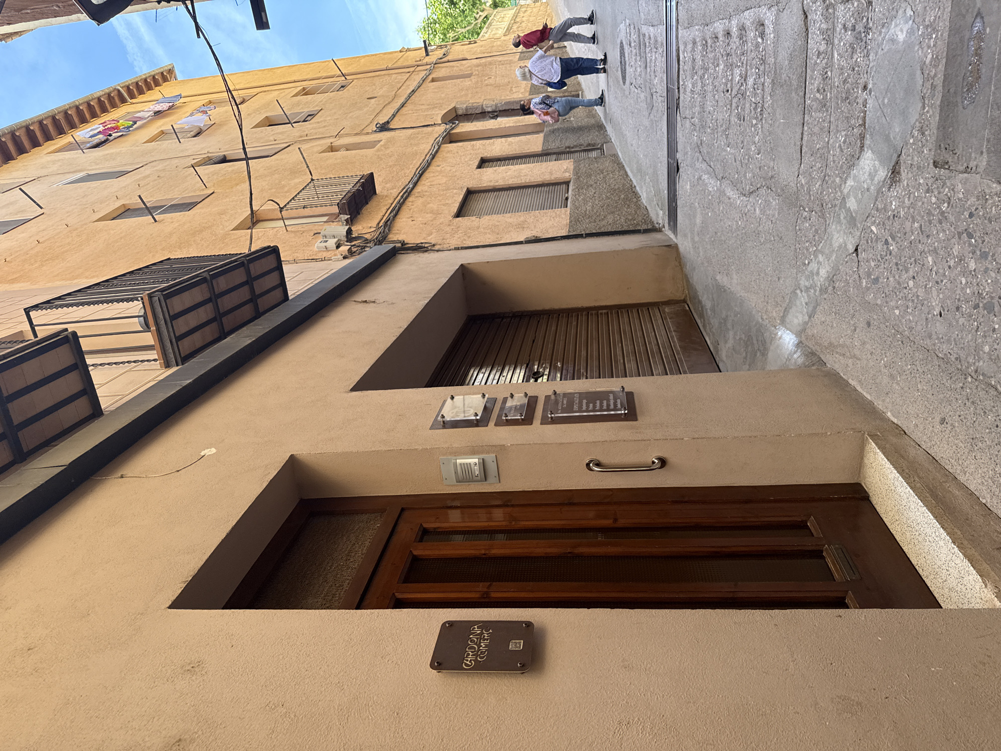The image shows a narrow street in the historic center of Cardona, Spain. The street is paved with uneven stone and concrete, with a slight incline. On the left is a building with a beige stucco facade, featuring a dark wooden door with glass panels and a bronze plaque that reads "CARDONA COMERC". To the right of the door, a metal handle is mounted on the wall, and several other plaques are visible, including one that appears to be a doorbell. Further down the street on the right, a multi-story building with an ochre-colored facade is visible. This building has several windows, balconies with wrought-iron railings, and laundry hanging from some of the upper floors. Three individuals are walking up the street on the right side of the image. The person in the foreground is a woman wearing a white shirt and dark pants, carrying a blue bag. Behind her, another woman is wearing a patterned shirt and dark pants, with a pink backpack. The third person, further ahead, is a man wearing a red jacket and gray pants. The sky above is blue with some scattered clouds.