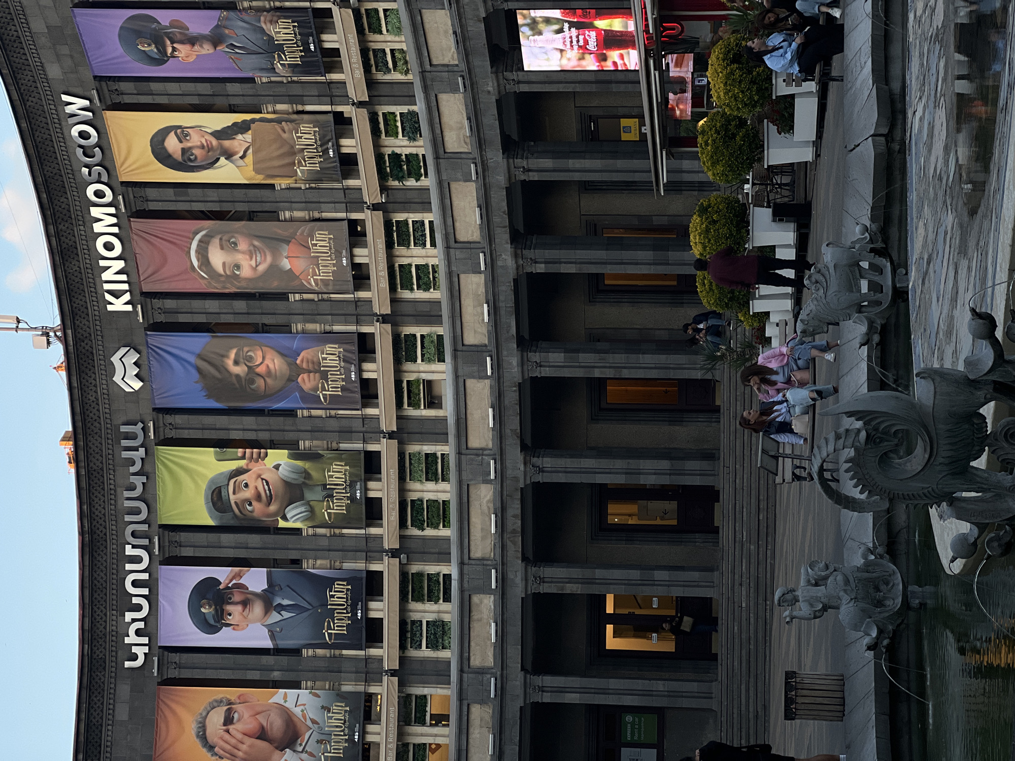 The image captures a scene at "KINOMOSCOW," as indicated by the large vertical sign on the building's facade, in an urban environment. The building displays a series of banners featuring cartoon characters, each accompanied by the text "Փոքր Սհեր" and the number "485." "Bar & Restaurant" signs are visible on the building as well.
People are seen near the building, some seated at tables, and others standing or walking by. The presence of a fountain with decorative sculptures adds to the scene. The environment includes a "UNIBANK" sign and an advertisement for "Coca-Cola," along with a sign reading "EXPRESS Rent a car."