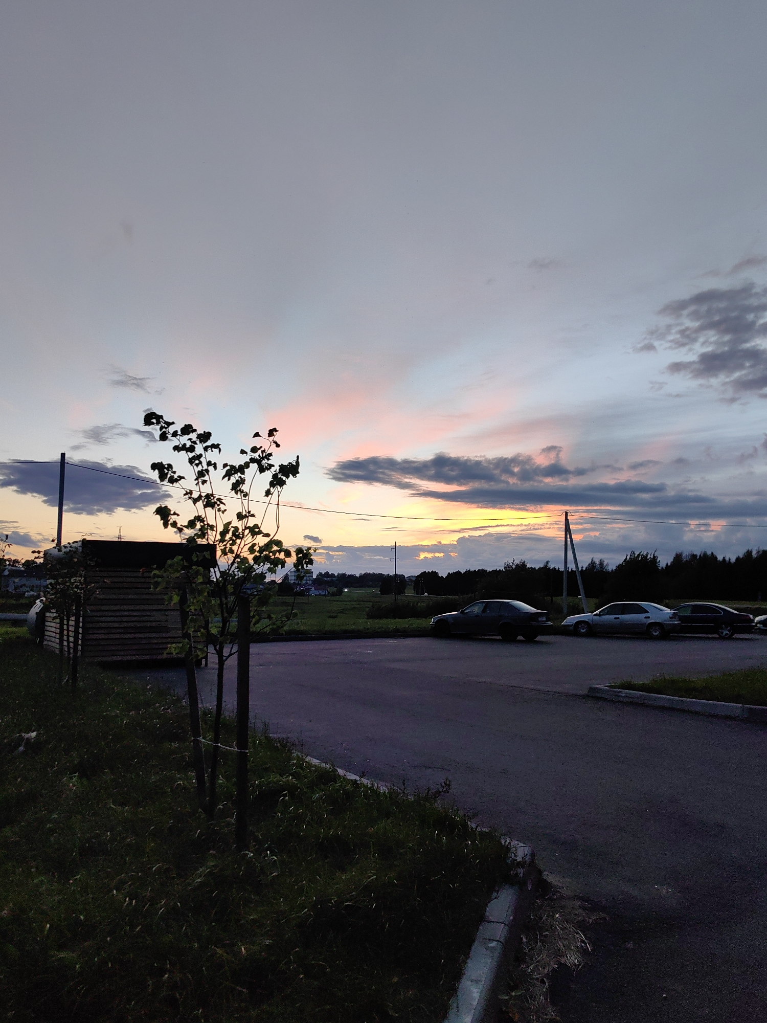 The image shows a rural roadside scene at dusk, possibly near Myadzyel, Belarus. A narrow asphalt road curves into the foreground, bordered by a strip of grass and a concrete curb. In the background, a line of dark trees forms the horizon, with a few scattered buildings and utility poles visible. Several cars are parked along the side of the road. To the left, a small wooden structure with a dark roof is partially visible, along with a young tree in the foreground. The sky is filled with dramatic clouds, painted in hues of pink, orange, and purple from the setting sun.