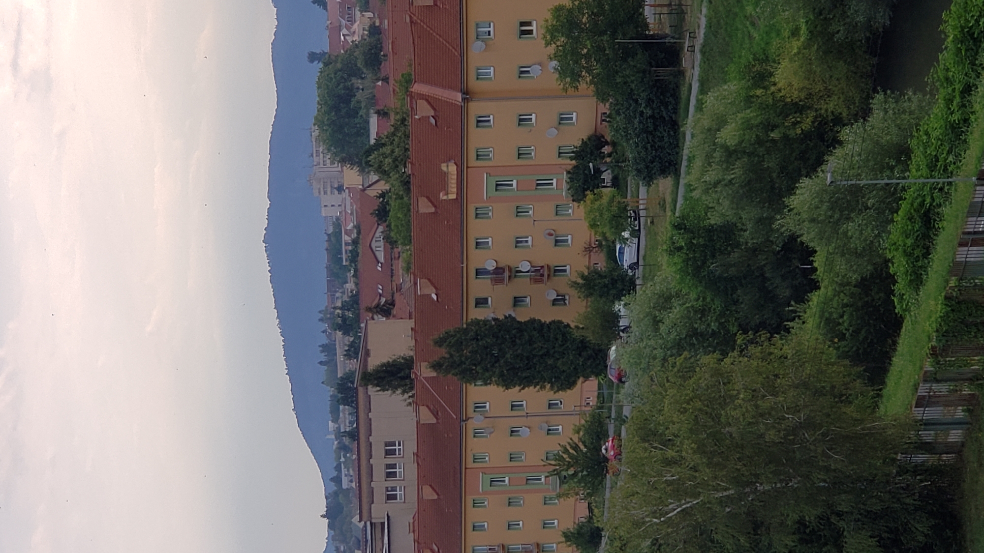 This is a medium-range, slightly elevated outdoor photograph of a residential area in Prešov, Slovakia. In the foreground, dense green foliage is visible, with a dark body of water to the right and a fence bordering a grassy area. Moving further into the frame, a long, multi-story apartment building with a yellow facade and red-tiled roof dominates the mid-ground. The building features numerous windows and satellite dishes. Adjacent to the building are trees of varying heights. Behind the apartment building, other residential structures with red roofs are visible, along with some taller buildings in the distance. A range of blue-tinted hills forms the backdrop under a hazy, overcast sky. Several cars are parked in front of the apartment building, and a street light is visible amongst the trees.